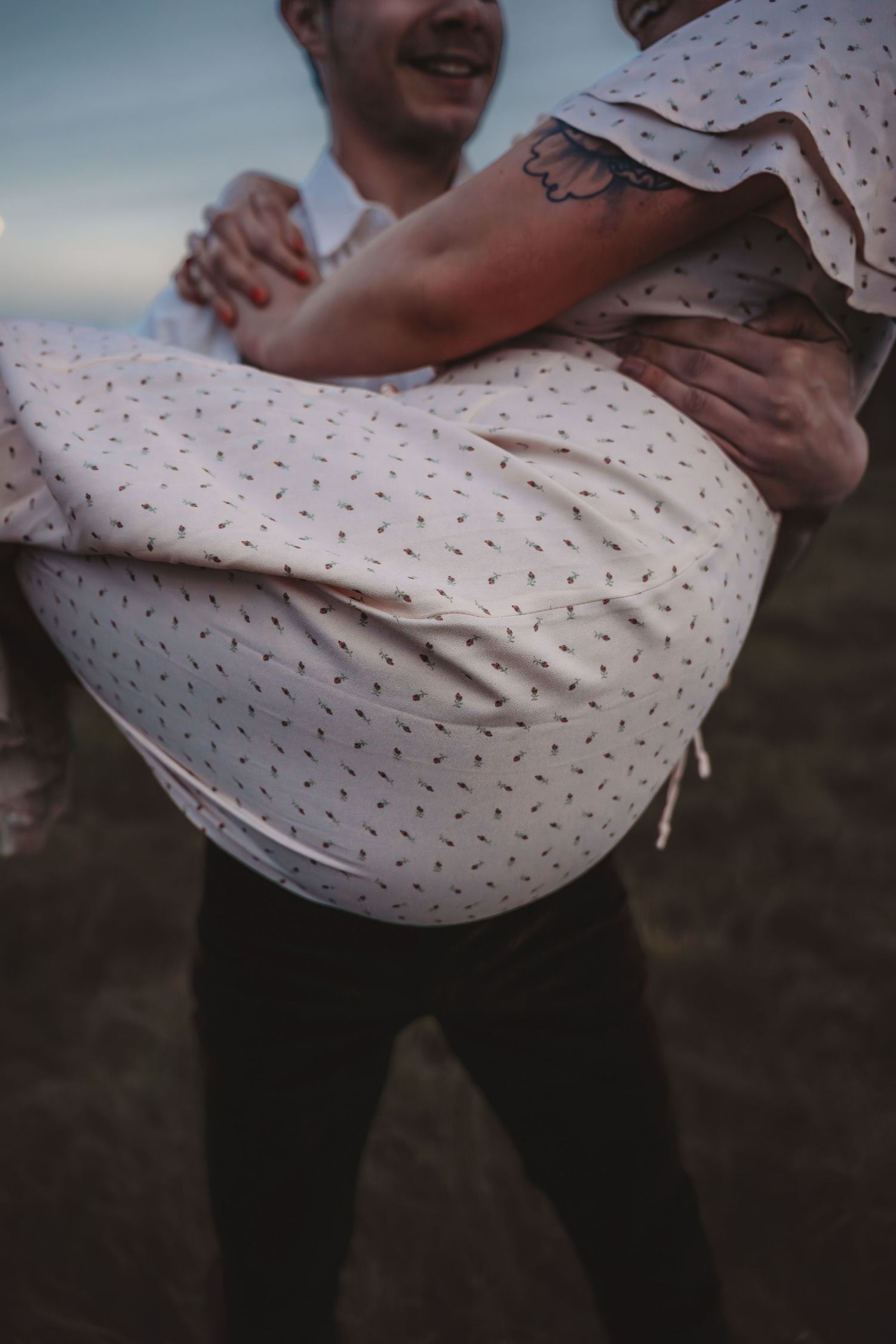 A man carries a woman in his arms outdoors, both smiling, she wears a floral dress and has a tattoo on her arm.