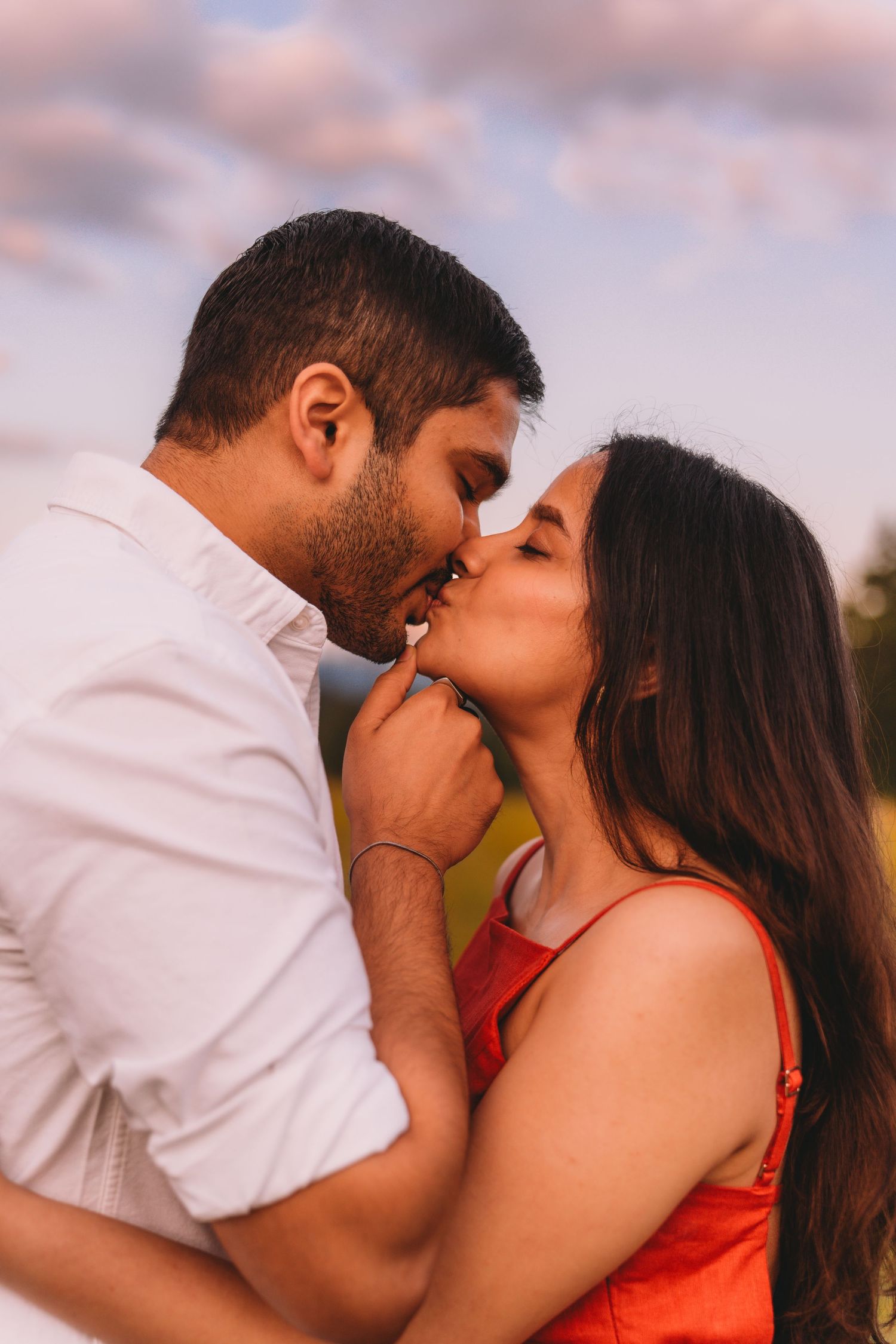 A romantic couple shares a tender kiss outdoors at golden hour, man in white shirt and woman in red dress.