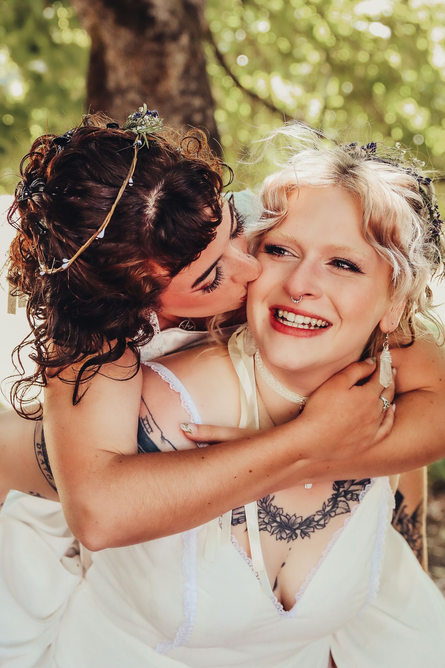 Two brides in white dresses laughing and embracing under a tree at their outdoor wedding celebration. Oregon