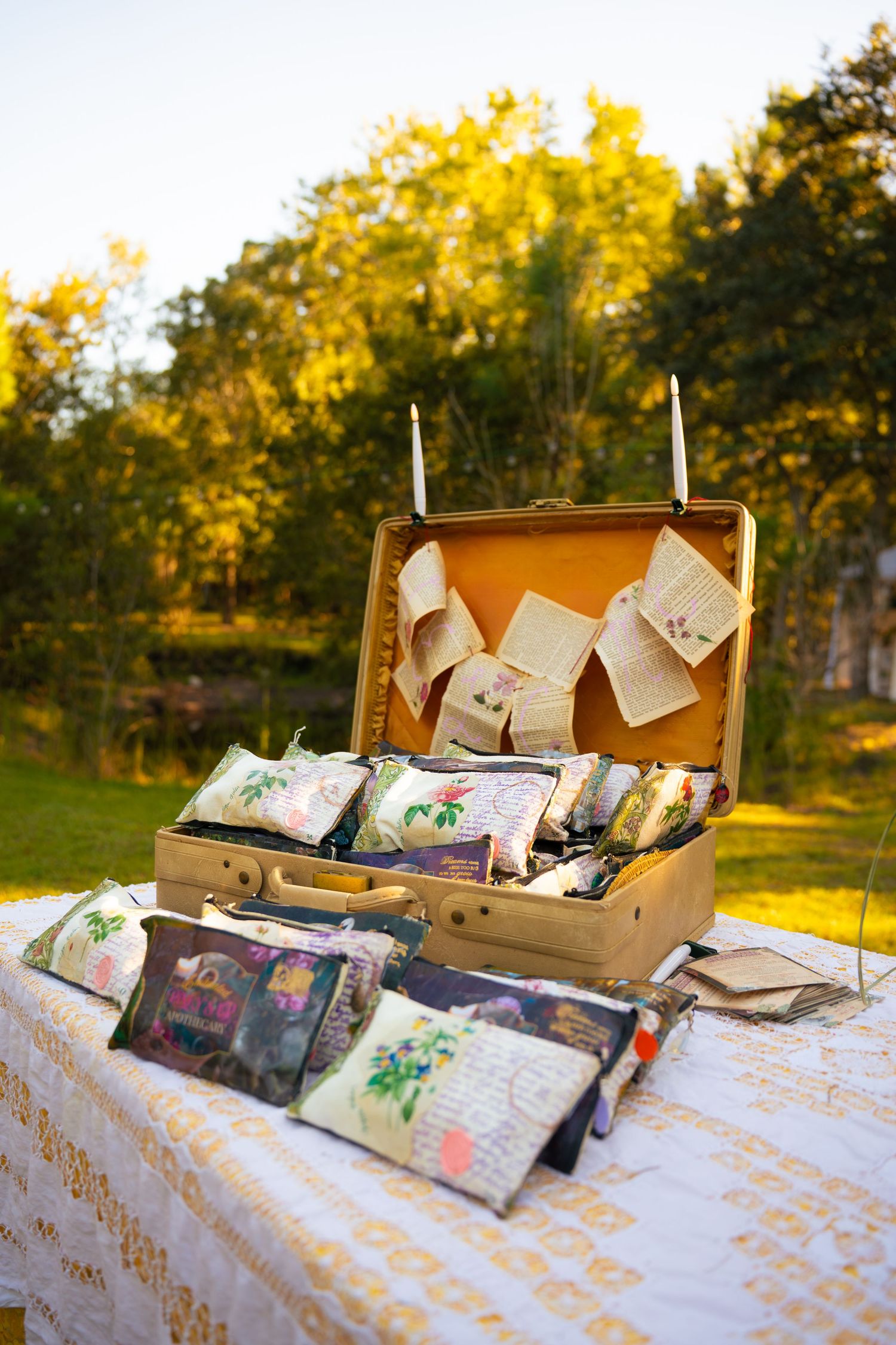 Vintage open suitcase filled with floral fabric sachets and handwritten notes on a lace-covered table outdoors among sunlit trees.