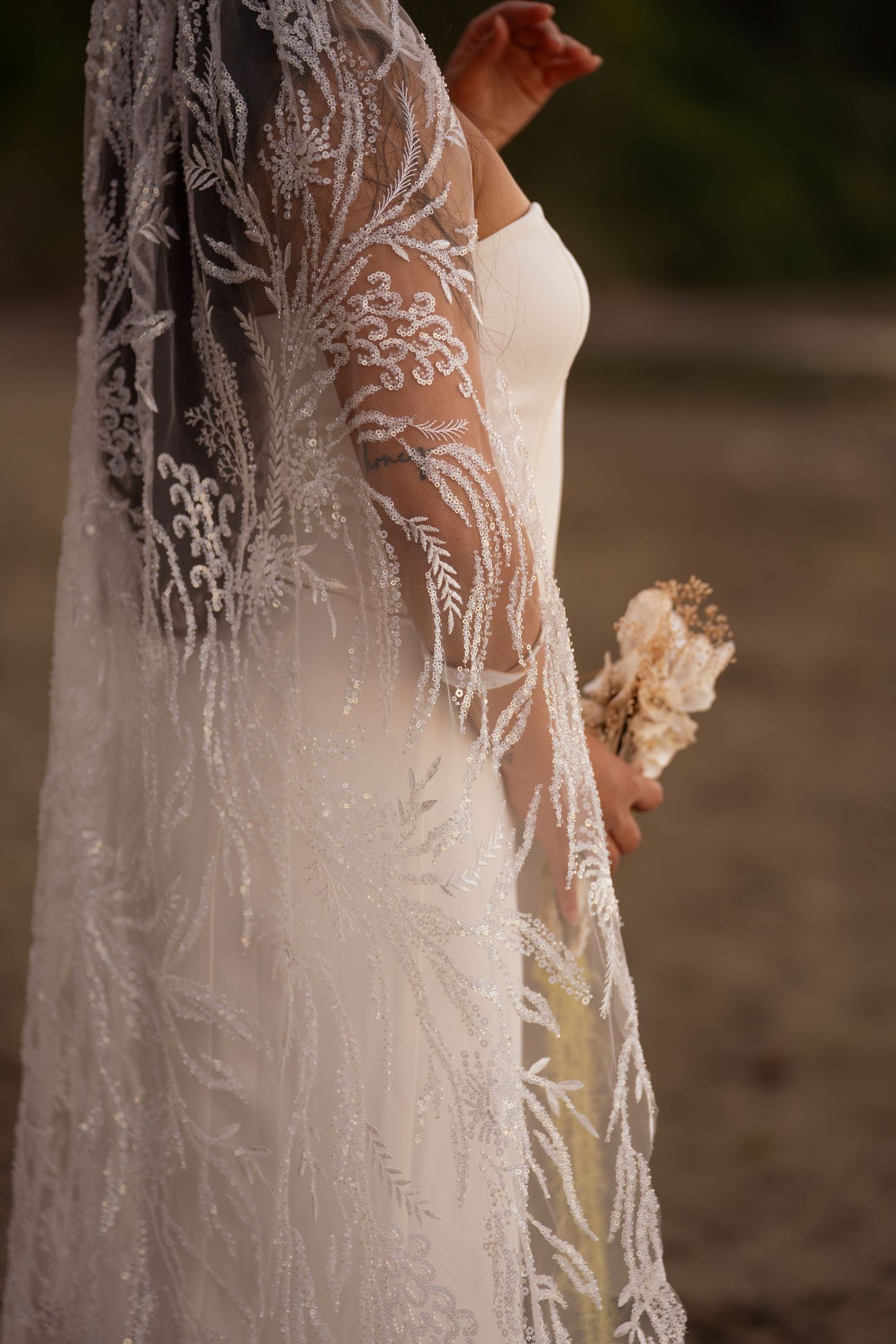 Bride in elegant beaded lace wedding veil and white gown holding dried floral bouquet outdoors.
