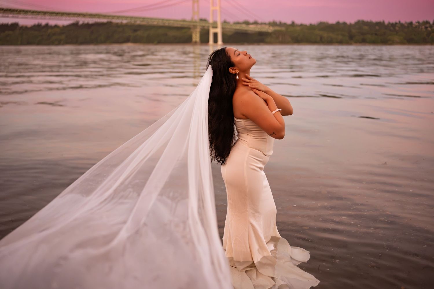 Bride in white gown and flowing veil poses gracefully by a river at sunset with a bridge in the background.