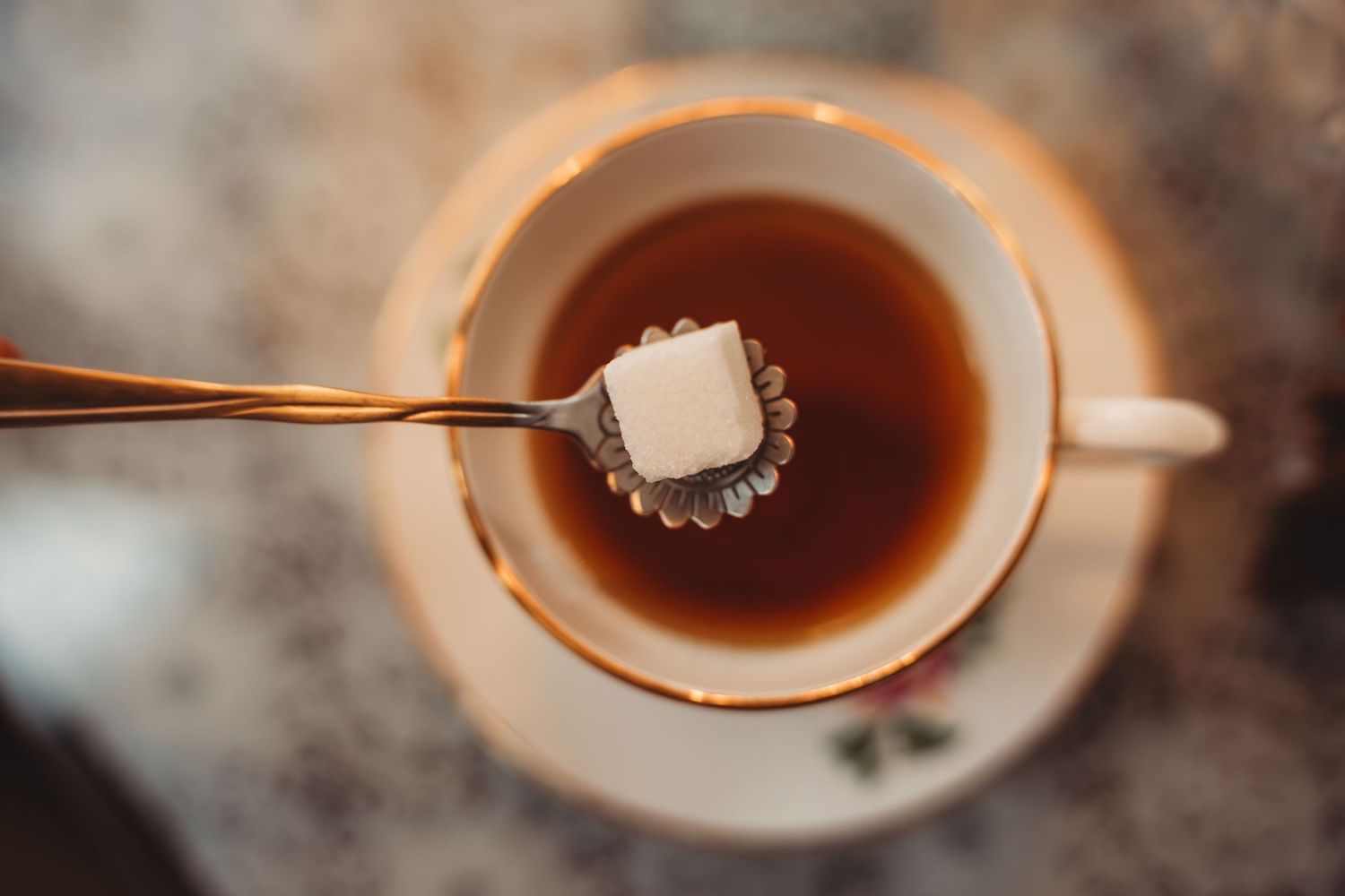 A silver spoon holding a sugar cube over a cup of hot tea on a saucer, elegant vintage china setting.
