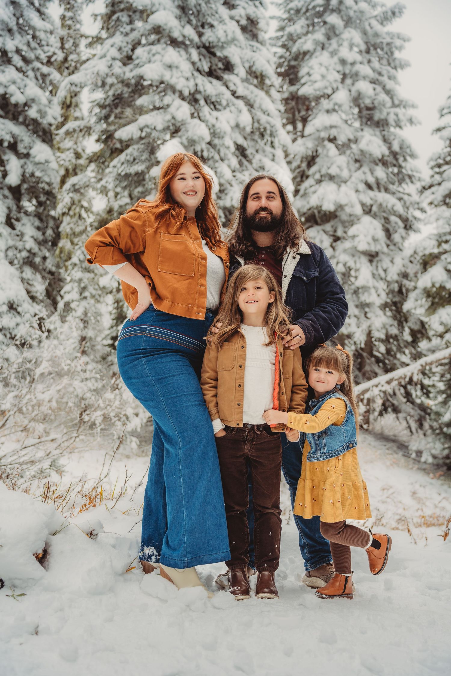 Family of four posing in snowy forest, parents and two girls in colorful winter outfits among snow-covered pine trees.