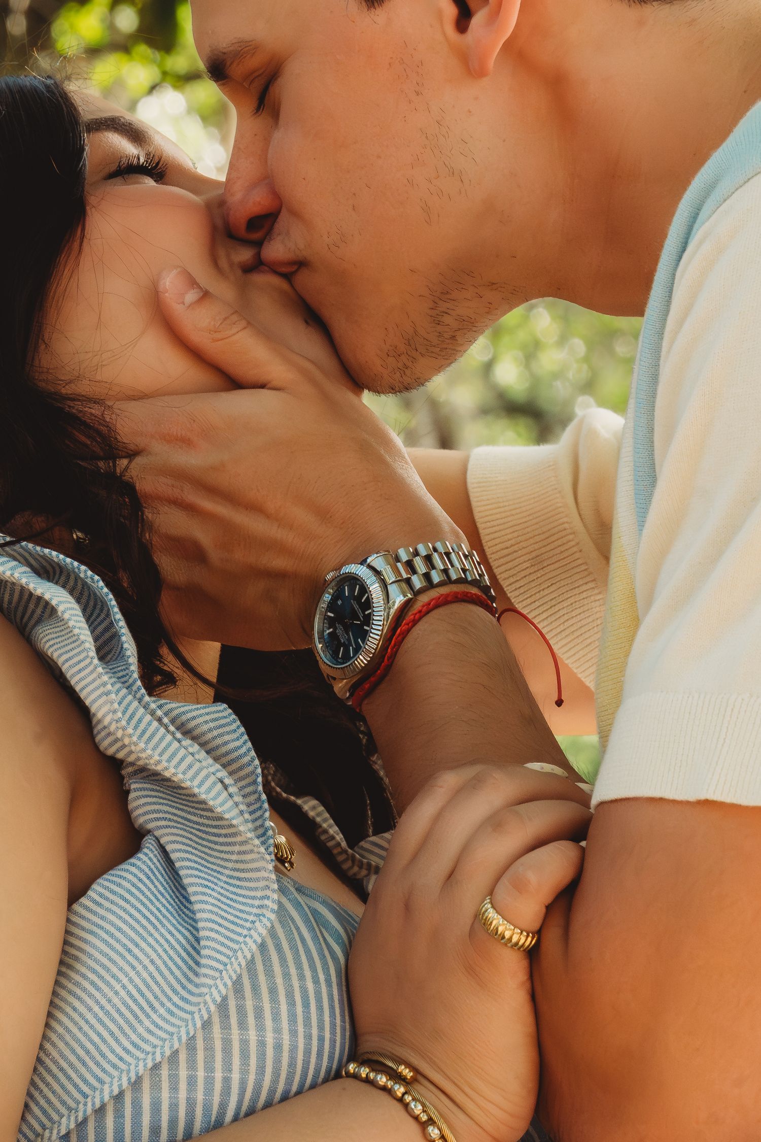 A couple sharing a romantic kiss outdoors, man wearing a silver watch and red bracelet, woman wearing gold jewelry.