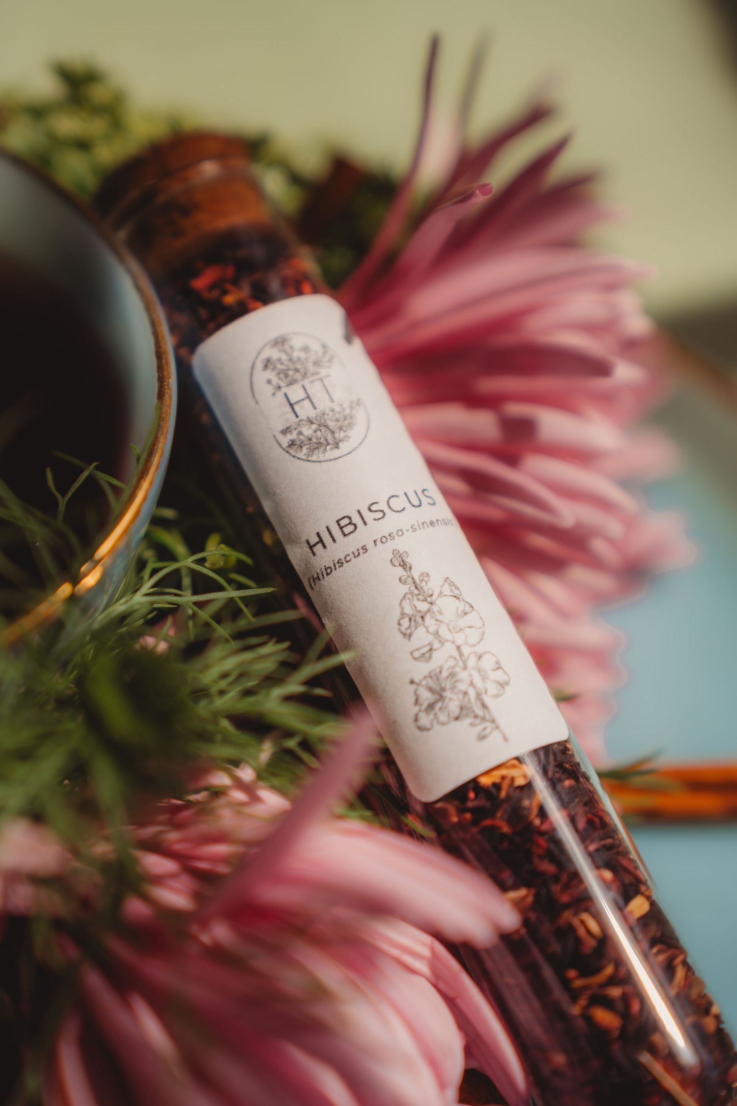 Glass tube of dried hibiscus tea with floral label surrounded by pink flowers.