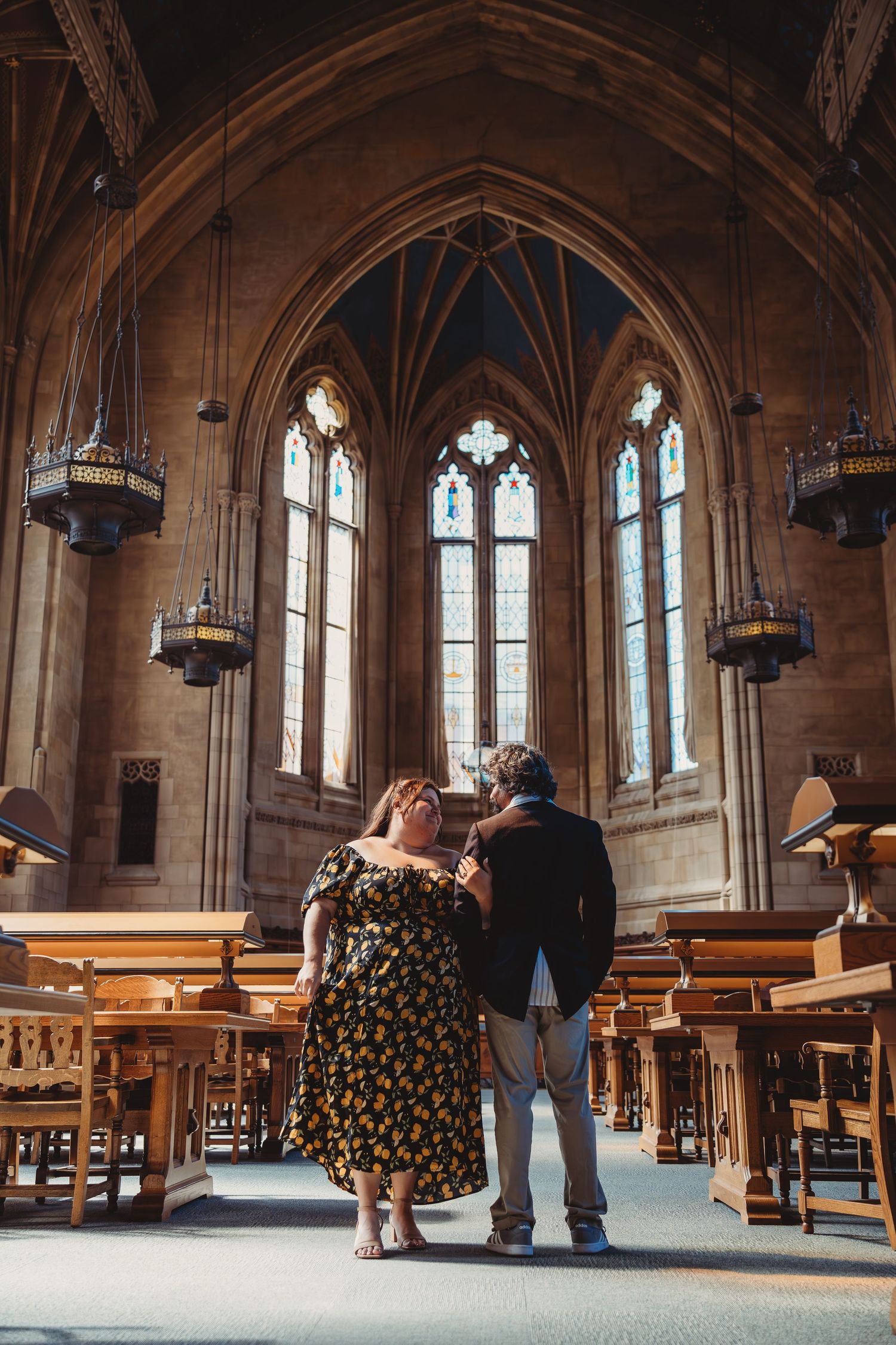 Couple embracing in a grand Gothic library with arched windows, wooden pews, and ornate stone architecture.