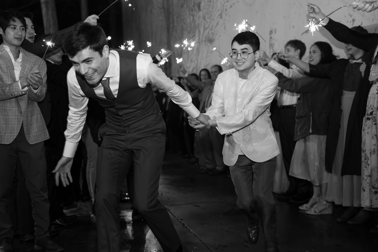 Two grooms hold hands running through sparkler exit at wedding reception in black and white photo.