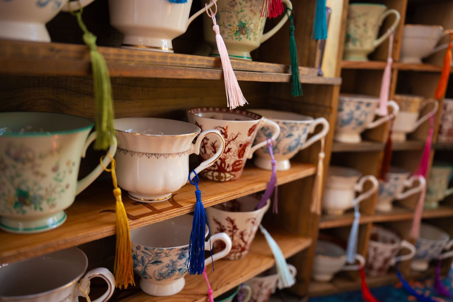 Vintage floral porcelain teacups with colorful silk tassels hanging from their handles on rustic wooden display shelves.
