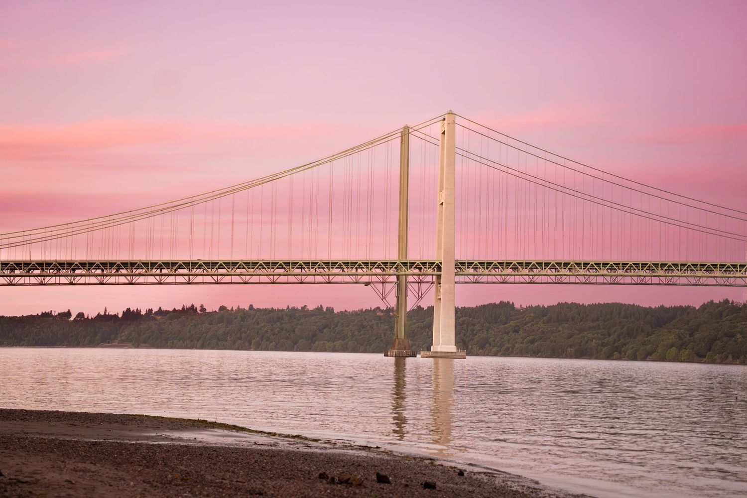 Suspension bridge over calm water at pink sunset with lush green trees in background.