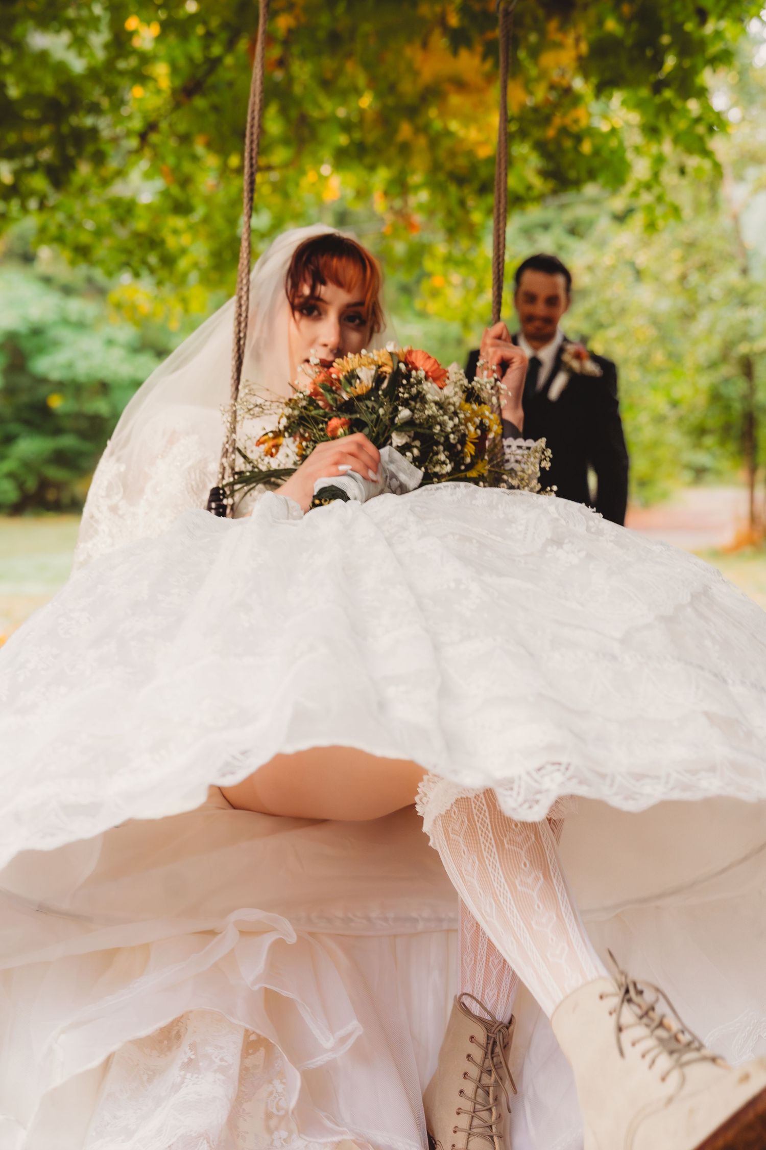 Bride in layered lace wedding dress sits on rope swing holding wildflower bouquet as groom stands in background.