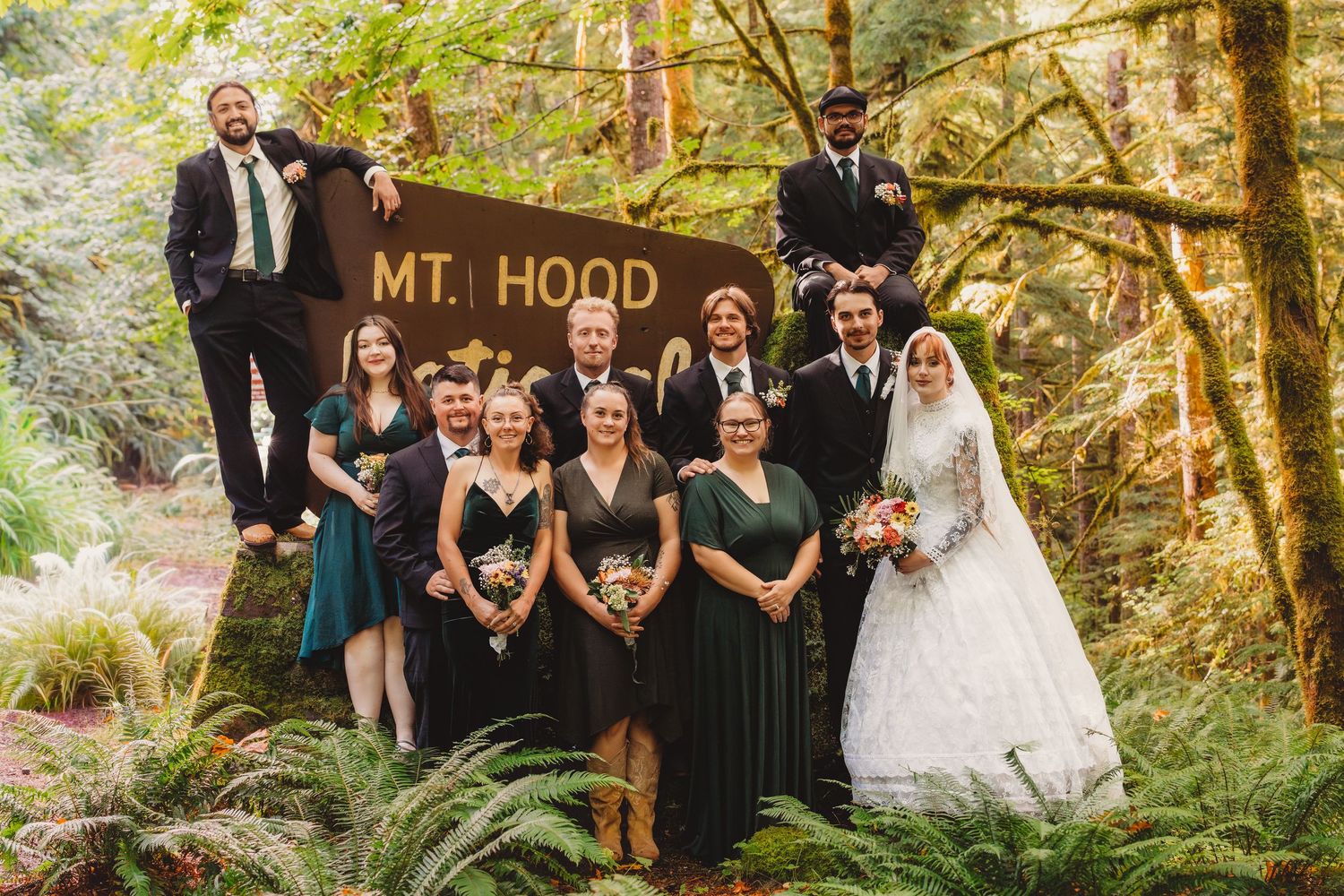 Wedding party posing by Mt. Hood sign in lush forest, bride in white gown with bridesmaids in green and black dresses.