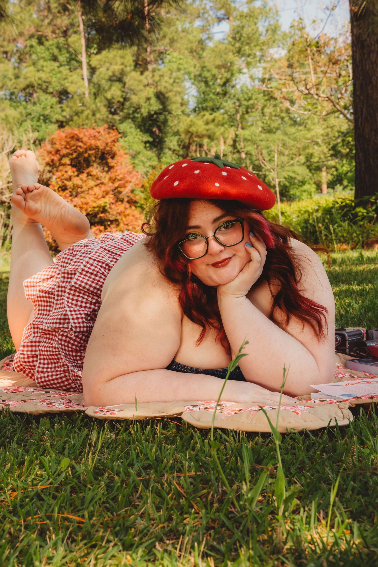 Plus-size woman in red gingham dress and strawberry beret relaxing on a picnic blanket in a lush green park.