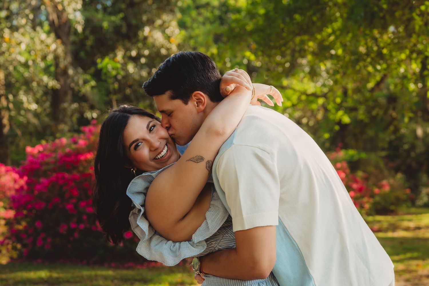 Couple embracing outdoors, man kissing woman's cheek as she smiles, with vibrant pink flowers and green trees behind.