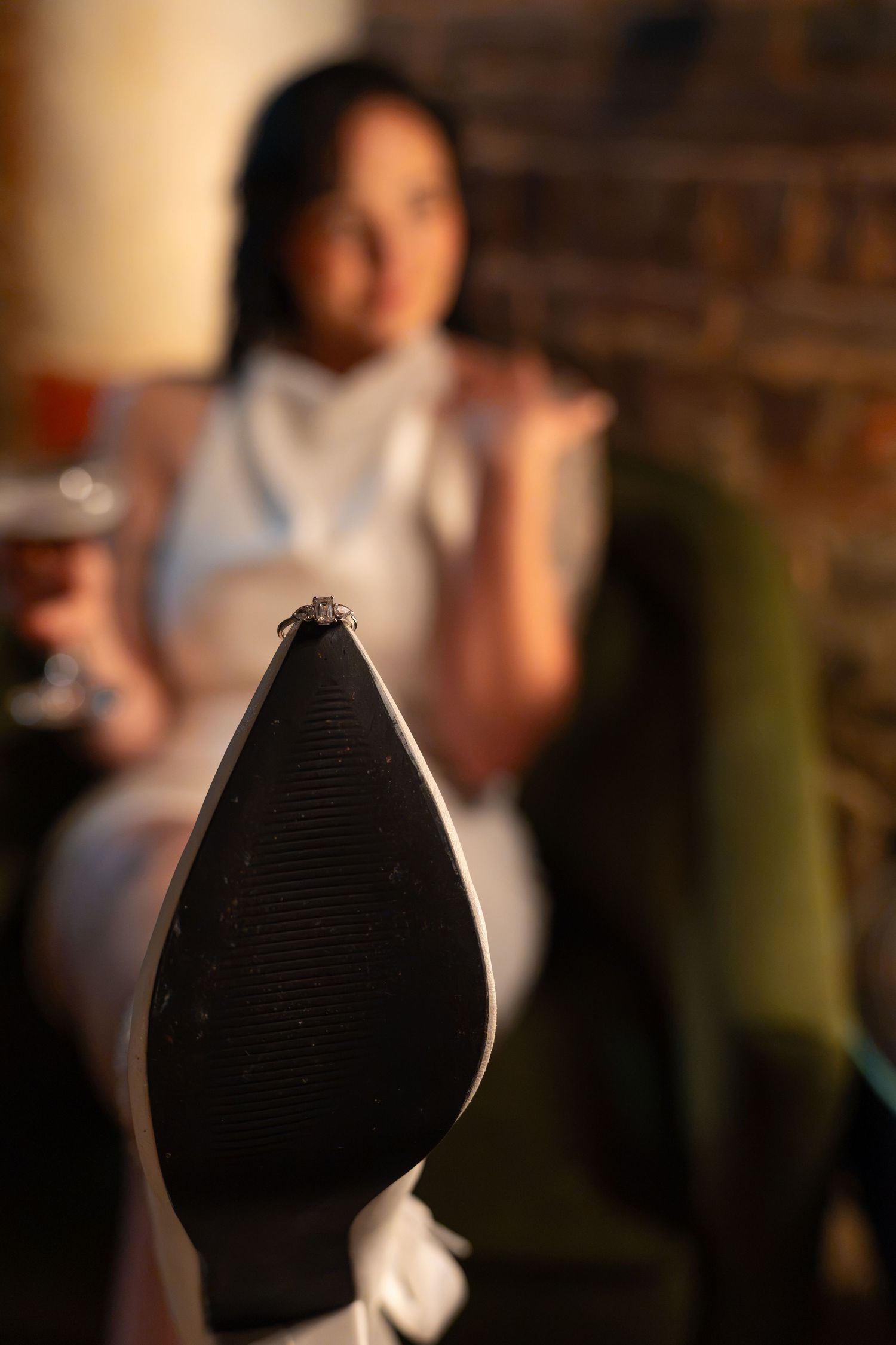 Sole of a white high heel with a diamond ring in the foreground and a blurred woman holding a drink behind it.