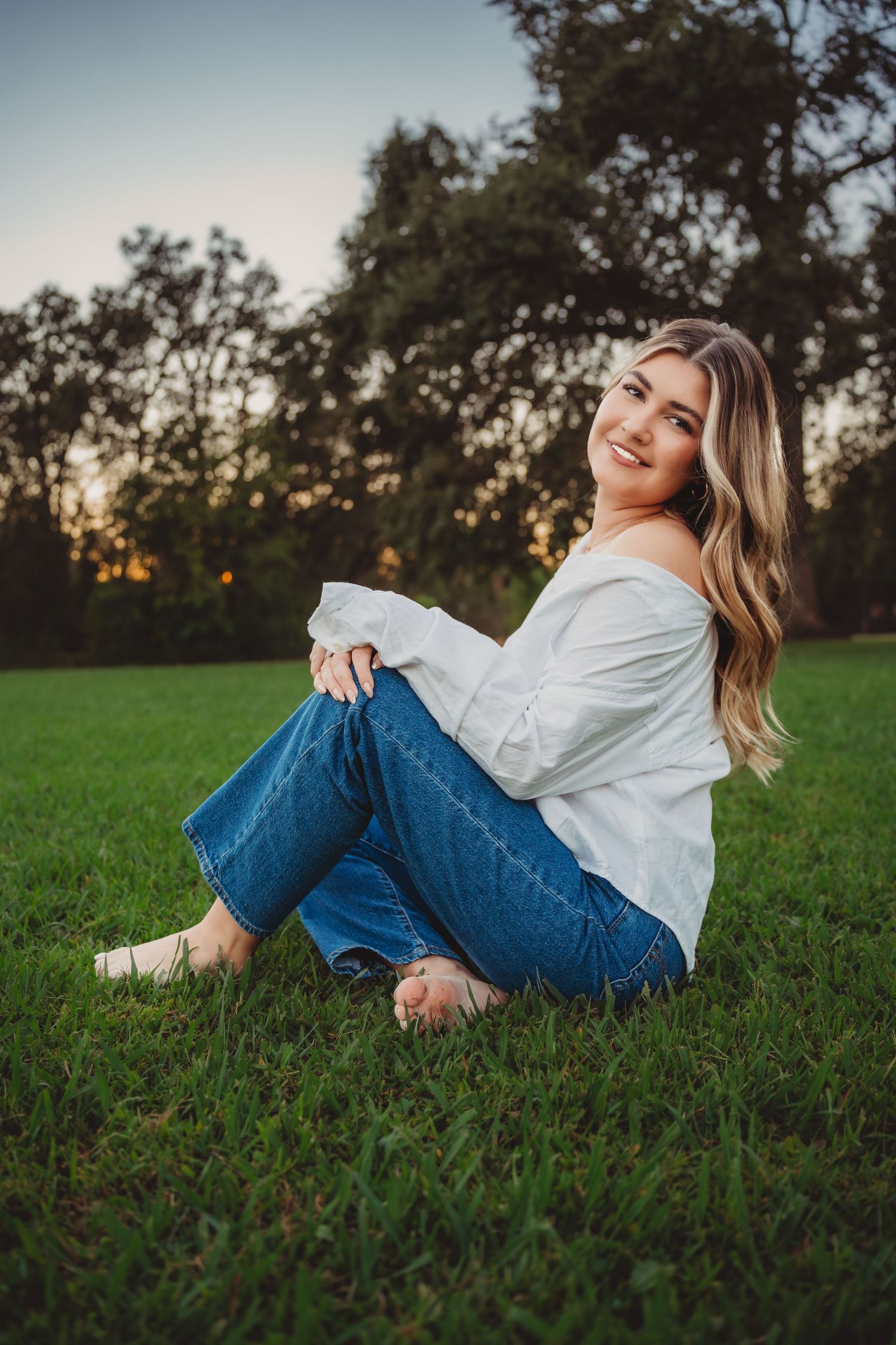 Smiling blonde teen girl sitting on green grass at sunset in white top and jeans.