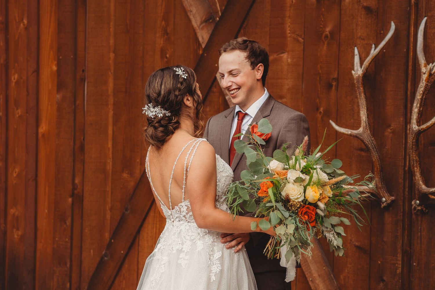 Smiling groom gazes at bride in backless lace gown before rustic wooden barn doors. Seattle