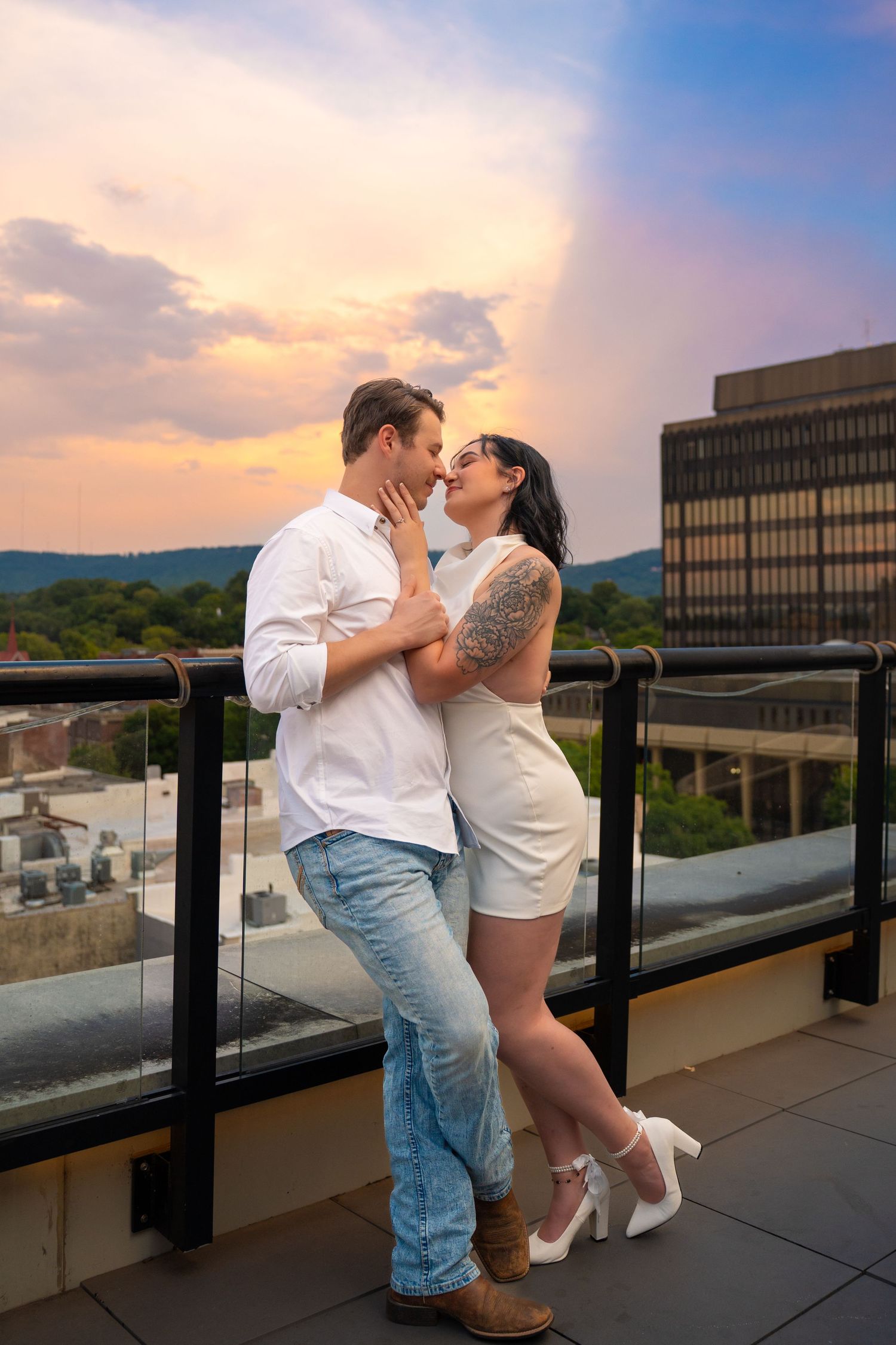 Couple embracing and kissing on a rooftop terrace at sunset with a glass railing, city buildings and hills in the background.