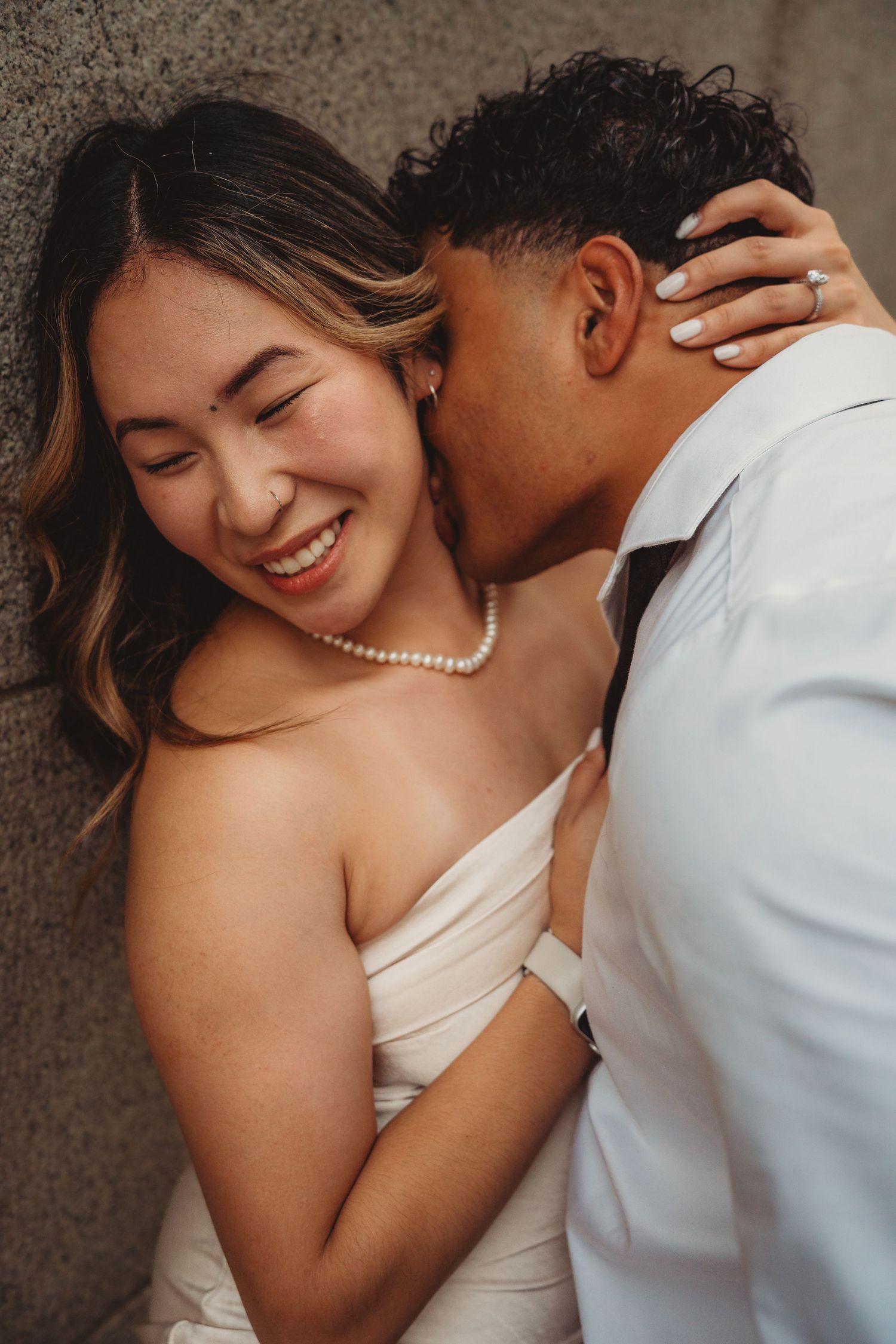 A joyful bride in a white strapless gown and pearl necklace laughs as her groom kisses her cheek.