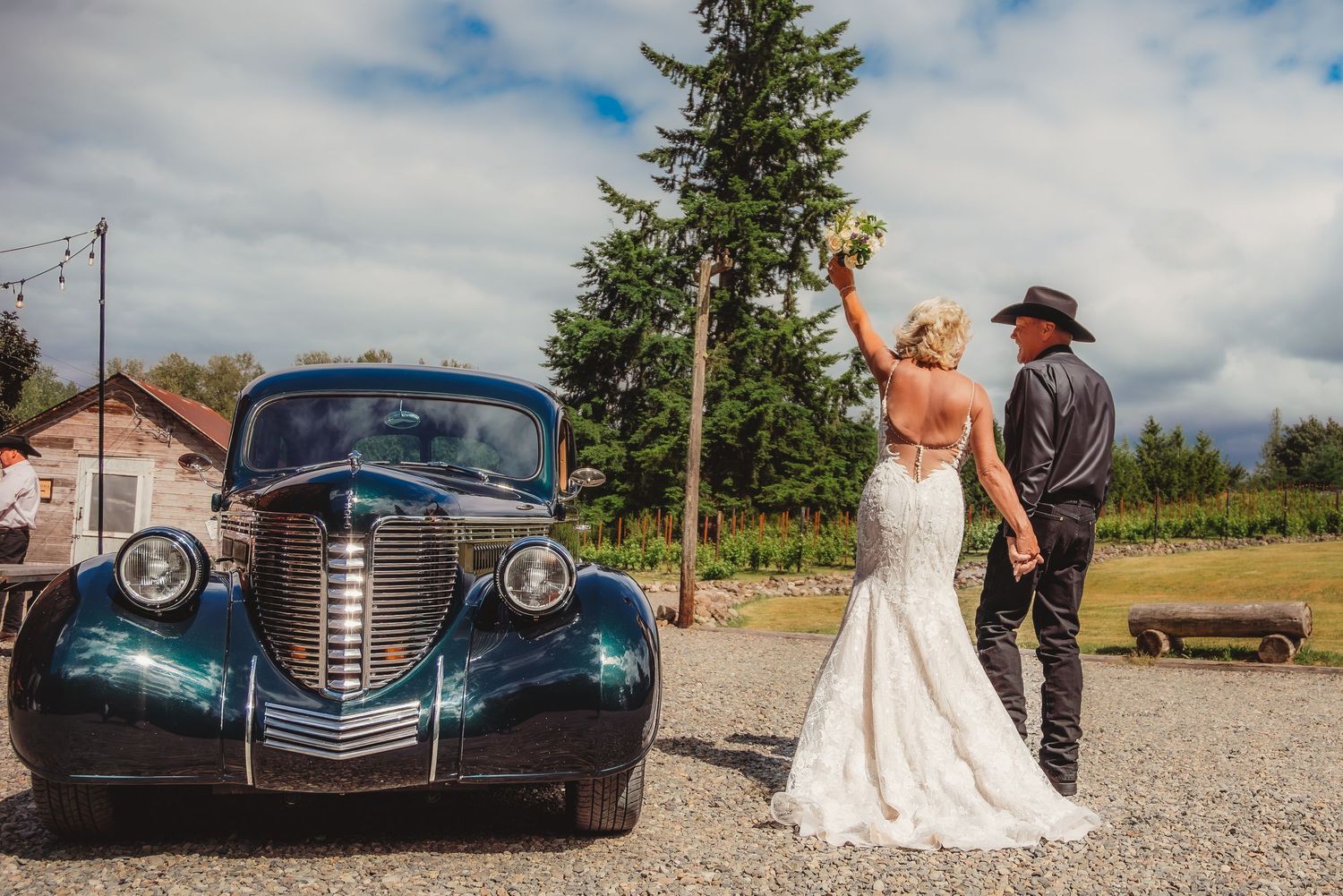 Bride in lace gown and groom in cowboy hat pose beside a vintage blue car at a rustic outdoor wedding venue.