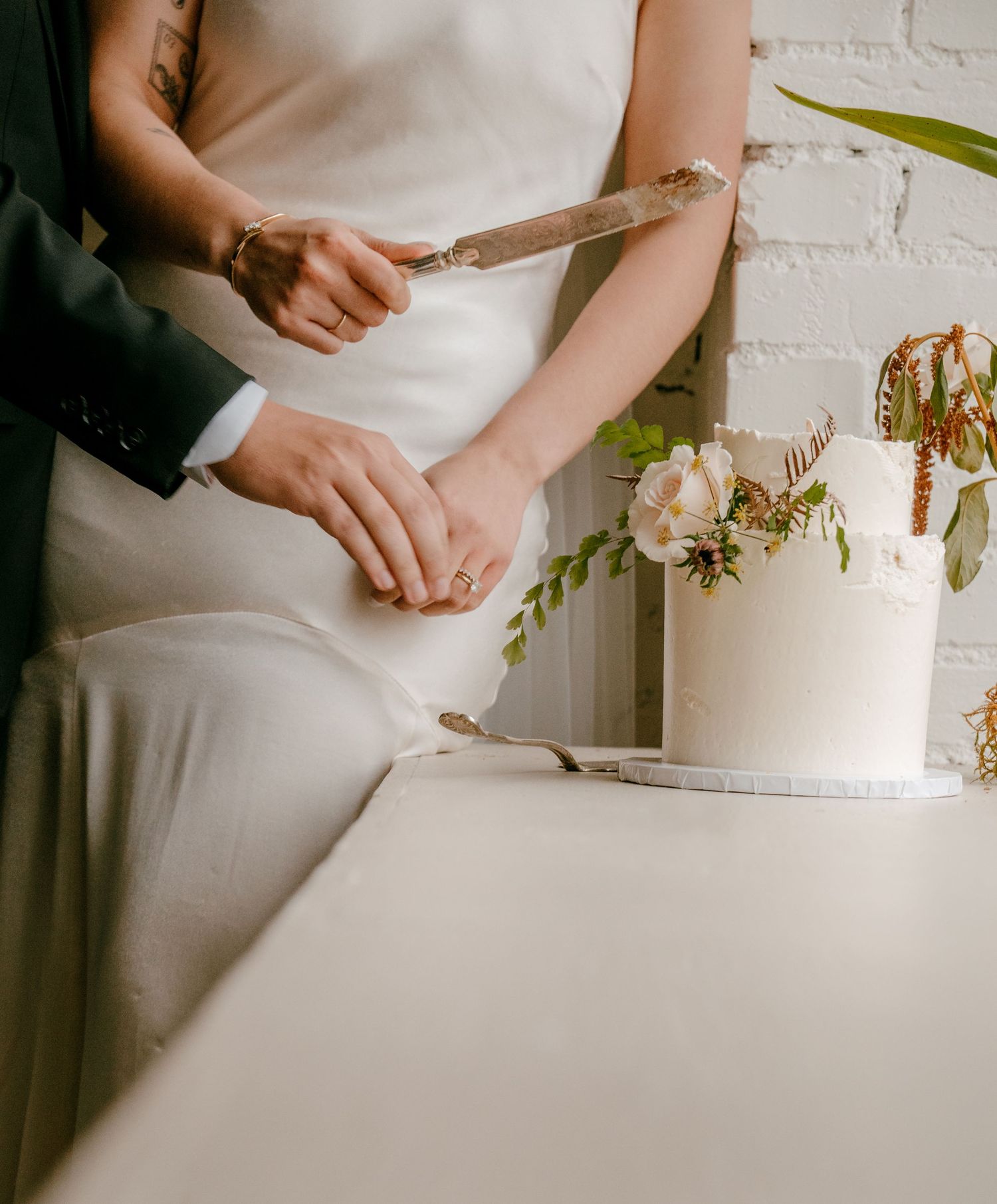 Bride and groom cutting a white wedding cake decorated with blush flowers and greenery at their reception.