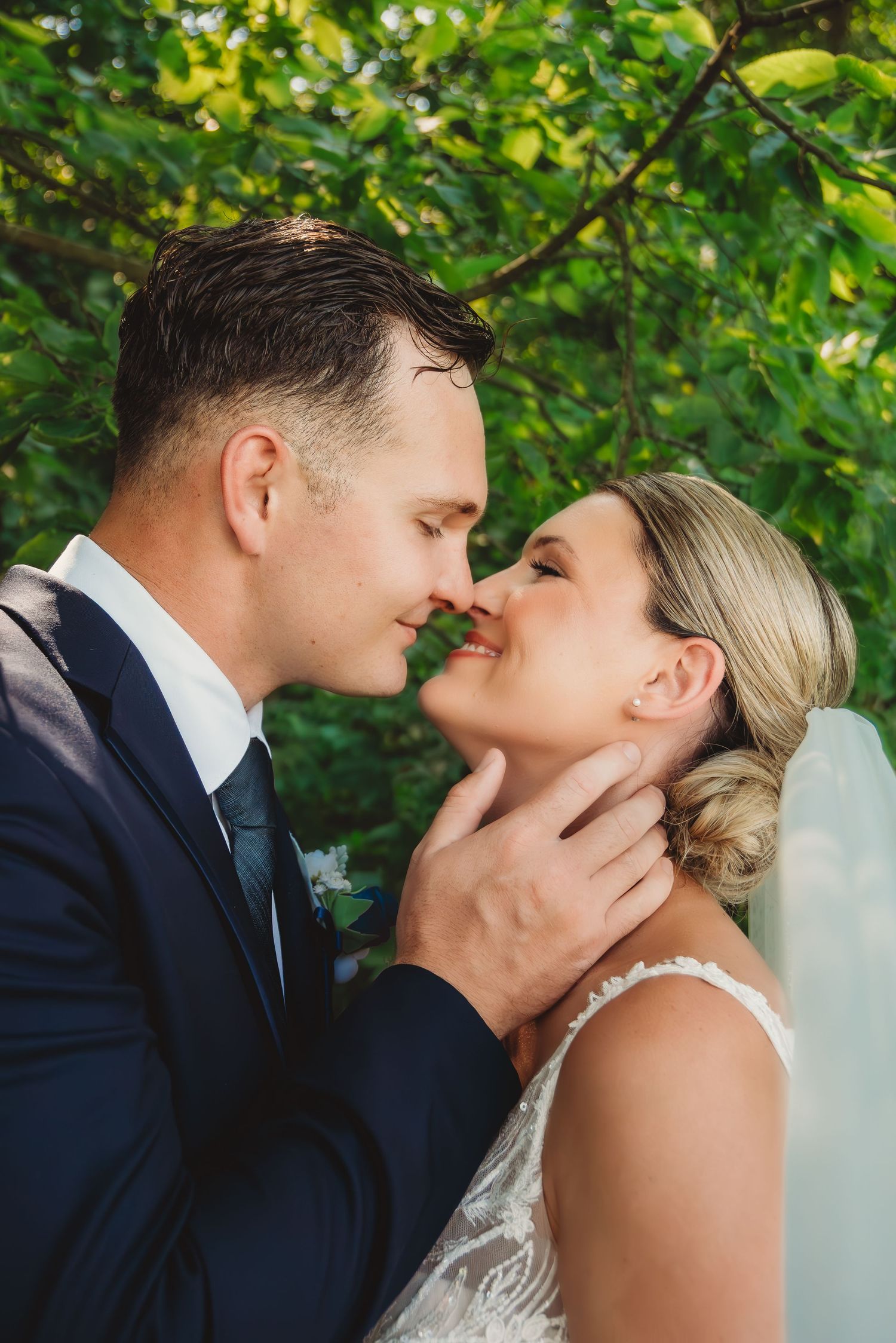 Groom tenderly holds brides face as they lean in for a kiss under green foliage. uptown Indiana