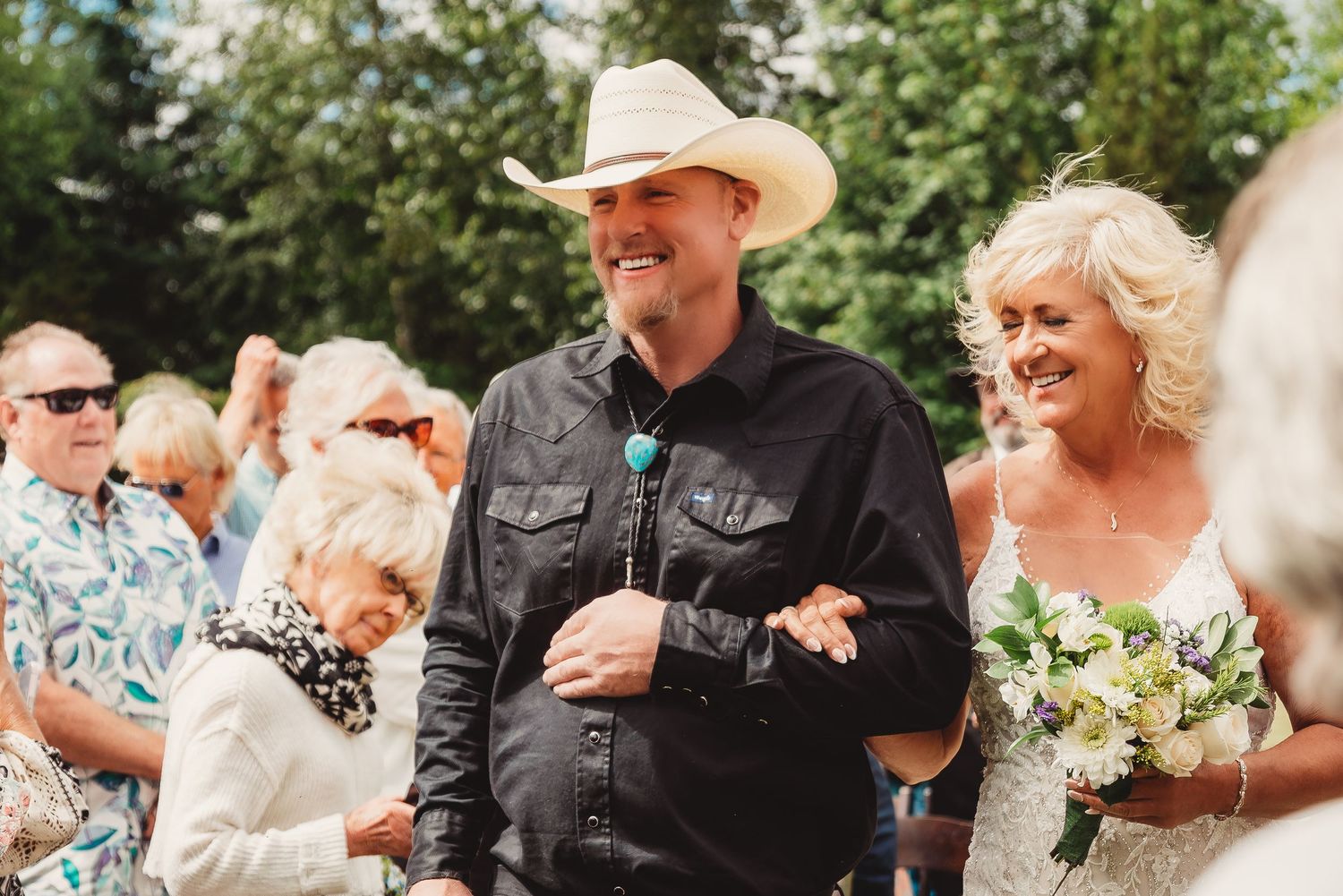 Smiling groom in cowboy hat and black shirt stands with blonde bride holding white bouquet at outdoor wedding ceremony.