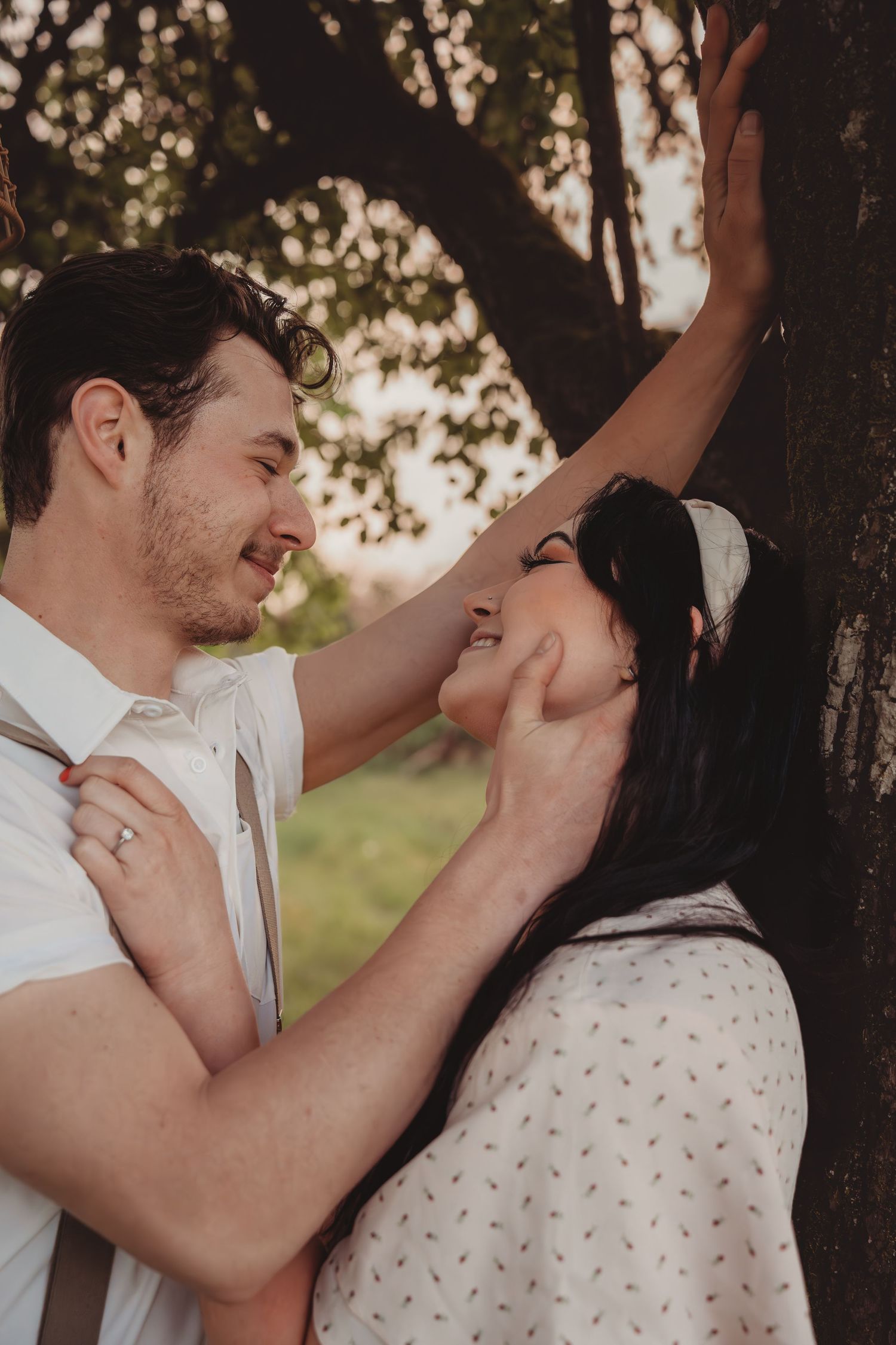 Romantic couple sharing a tender moment by a tree in a sunlit park, man gently holding woman's face.