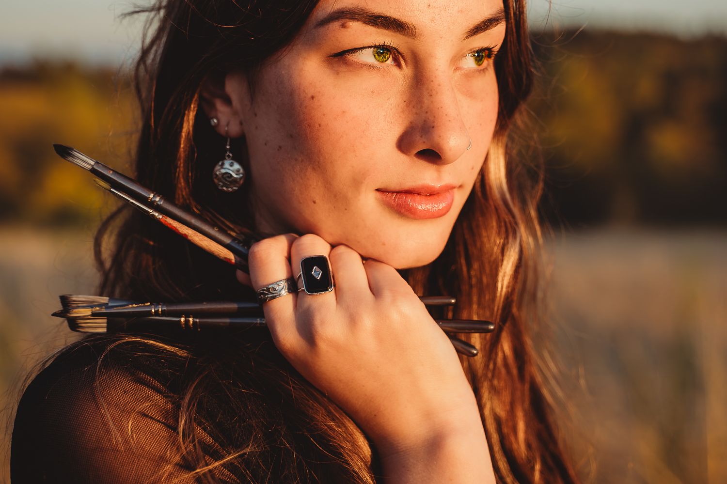 Young woman artist holding paint brushes near her face, wearing a black ring, with golden autumn bokeh background.
