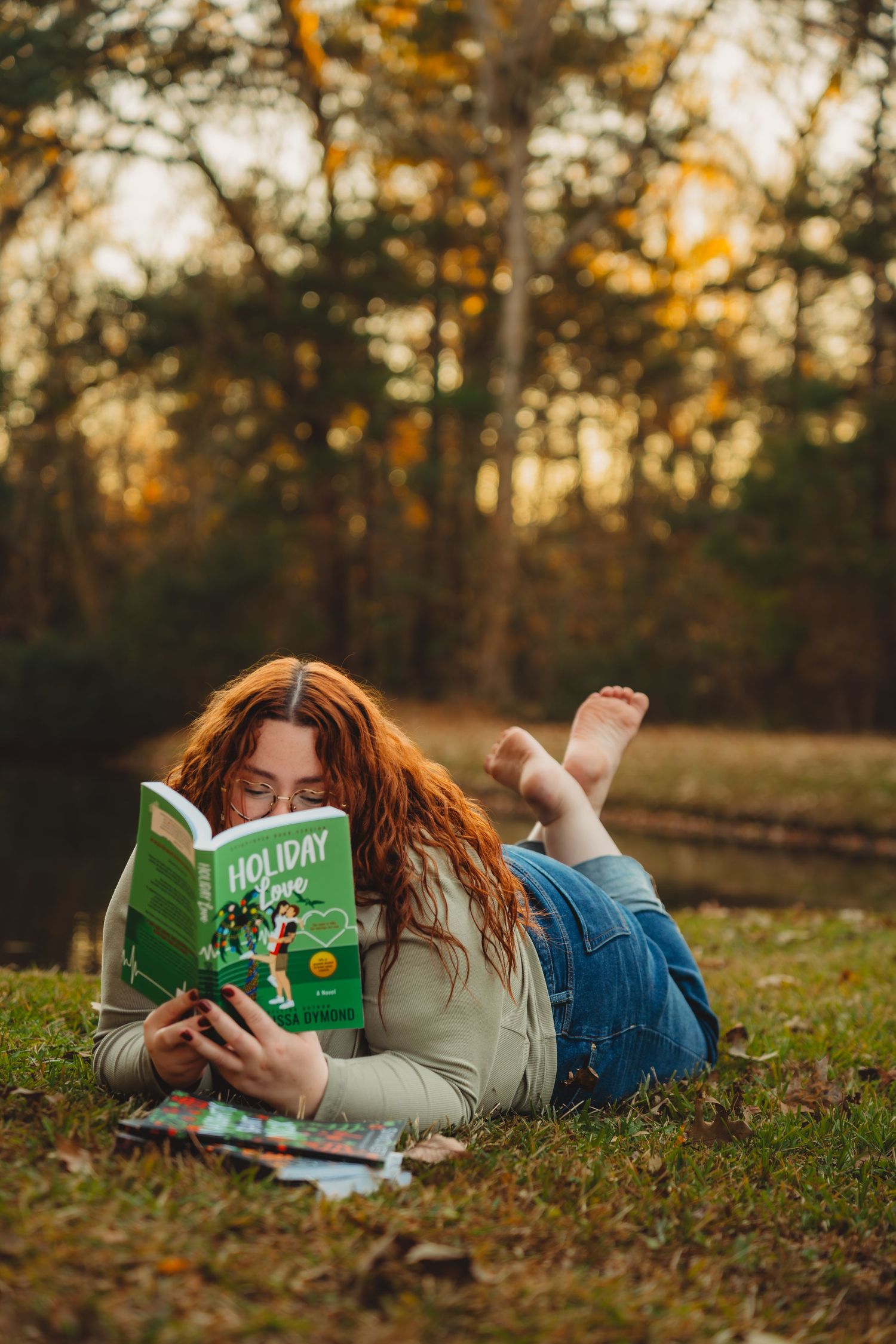 Red-haired woman reading Holiday Love book while lying on grass in autumn park.