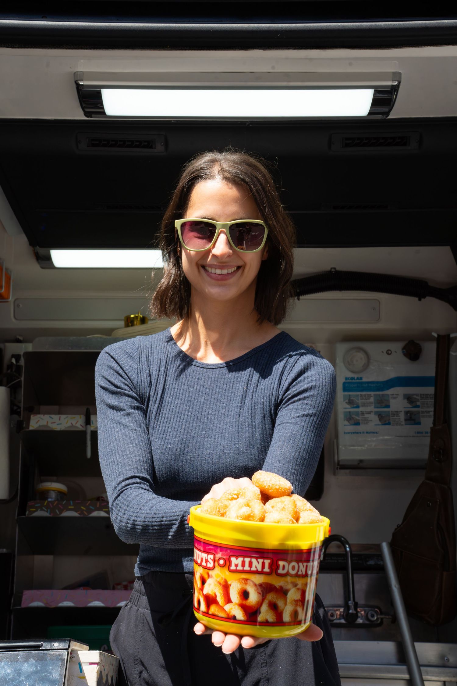 Smiling woman in sunglasses holding a bucket of mini donuts in front of a food truck. West Ashley South Carolina