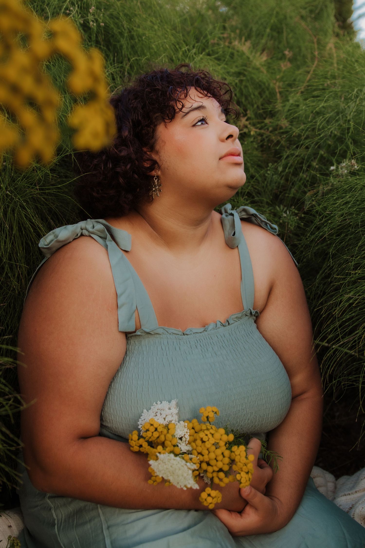 Plus size woman in blue sundress holding yellow wildflowers while sitting in tall green grass outdoors.Charleston South Carolina