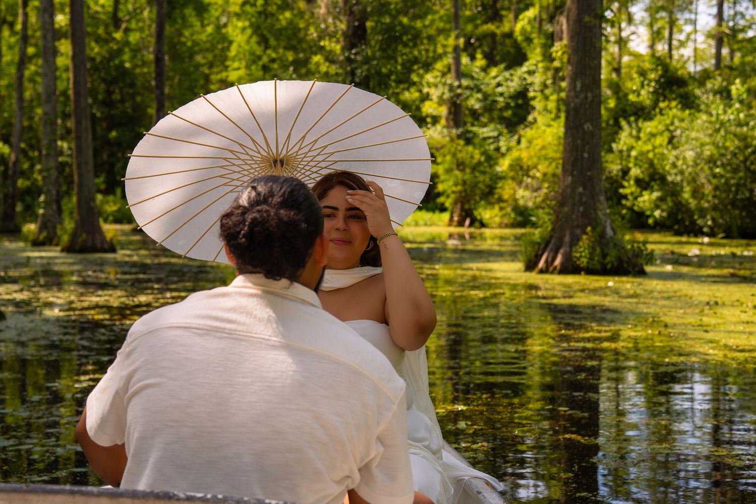 Couple in white attire sharing romantic moment on swamp boat, woman holding parasol umbrella amid cypress trees.