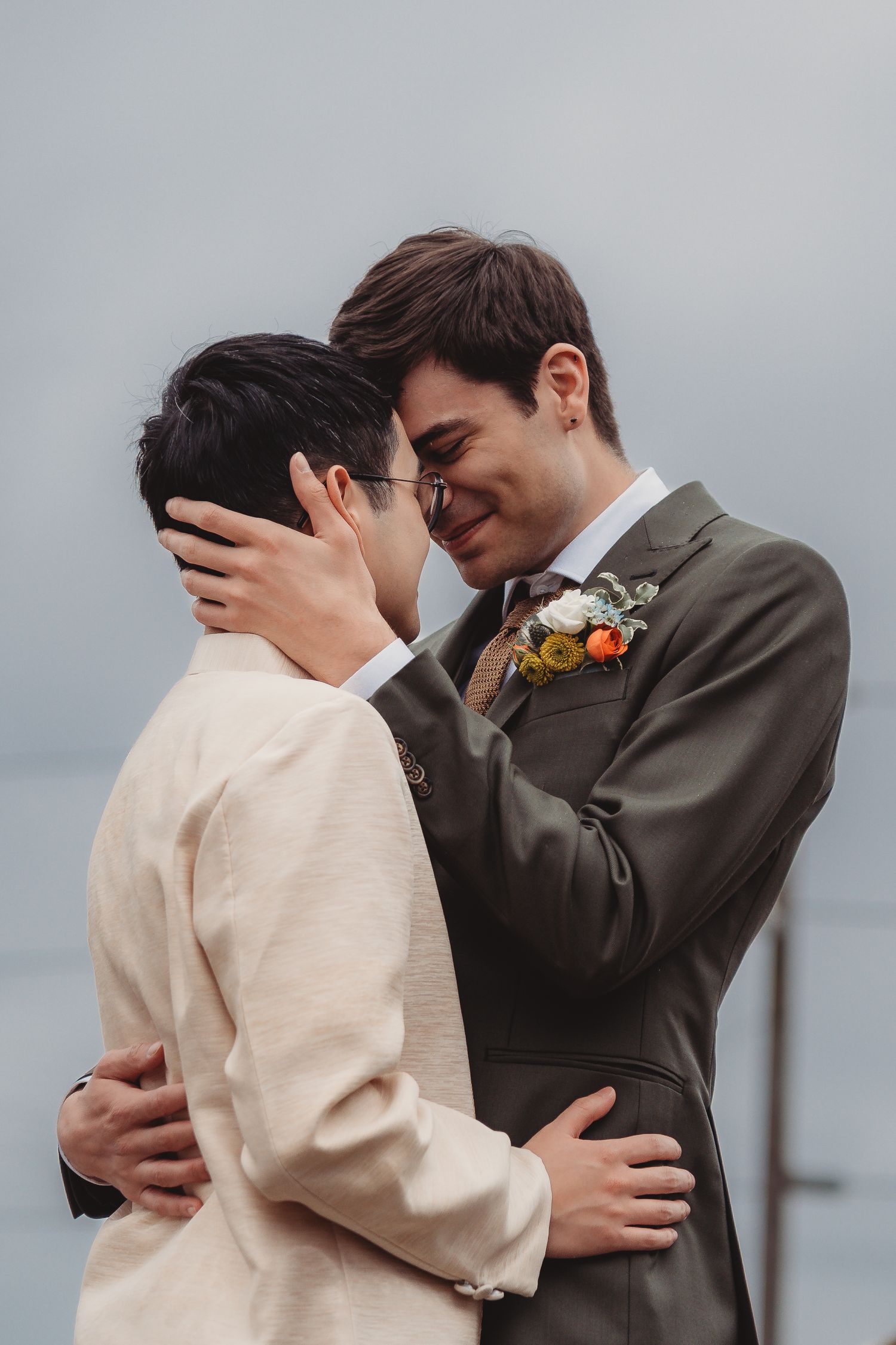 Two grooms in suits share an intimate moment, one wearing a floral boutonniere, at an outdoor wedding ceremony. Seattle