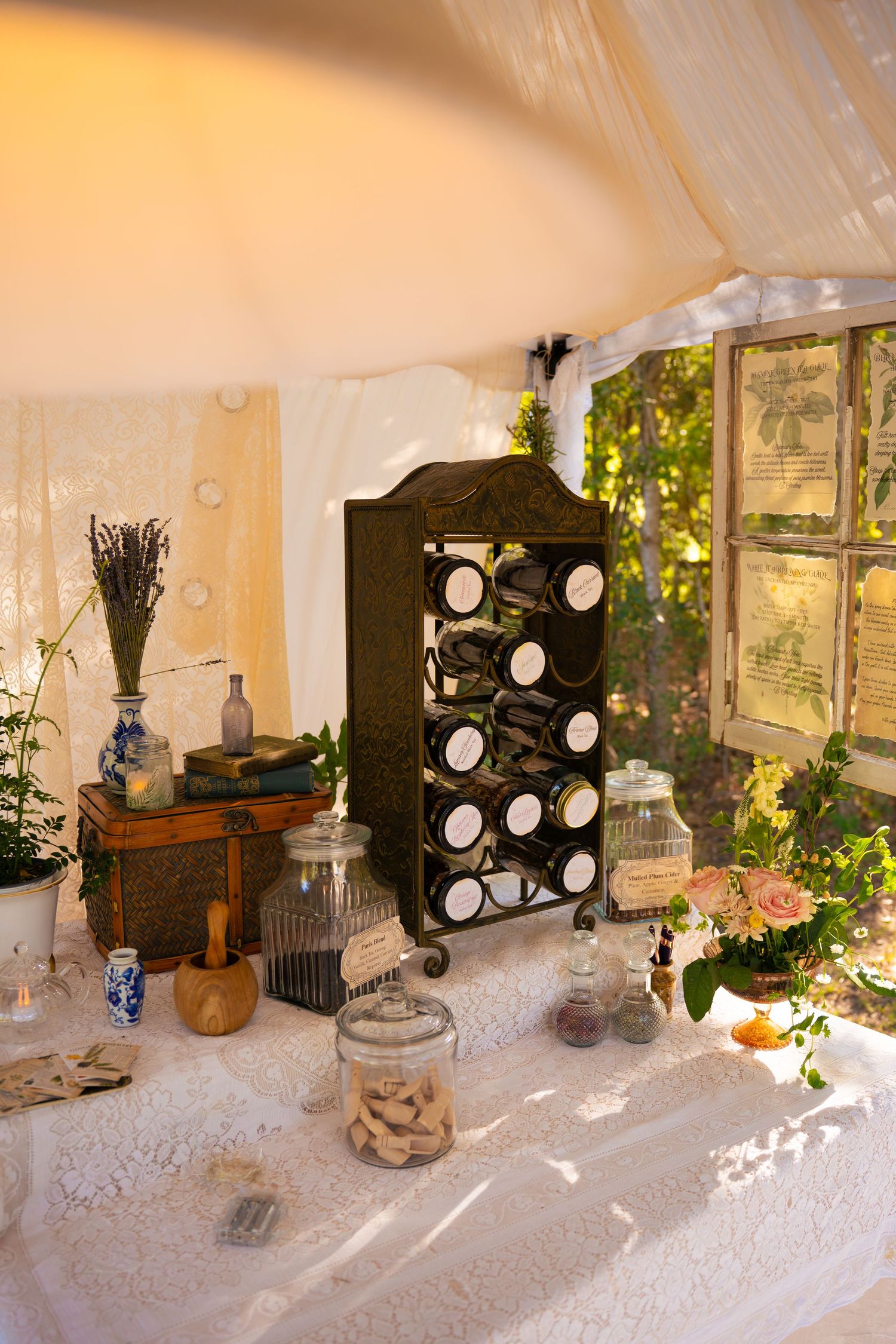 Rustic wedding table with a black spice rack, glass apothecary jars, lavender and pink roses on white lace under a sheer canopy tent.