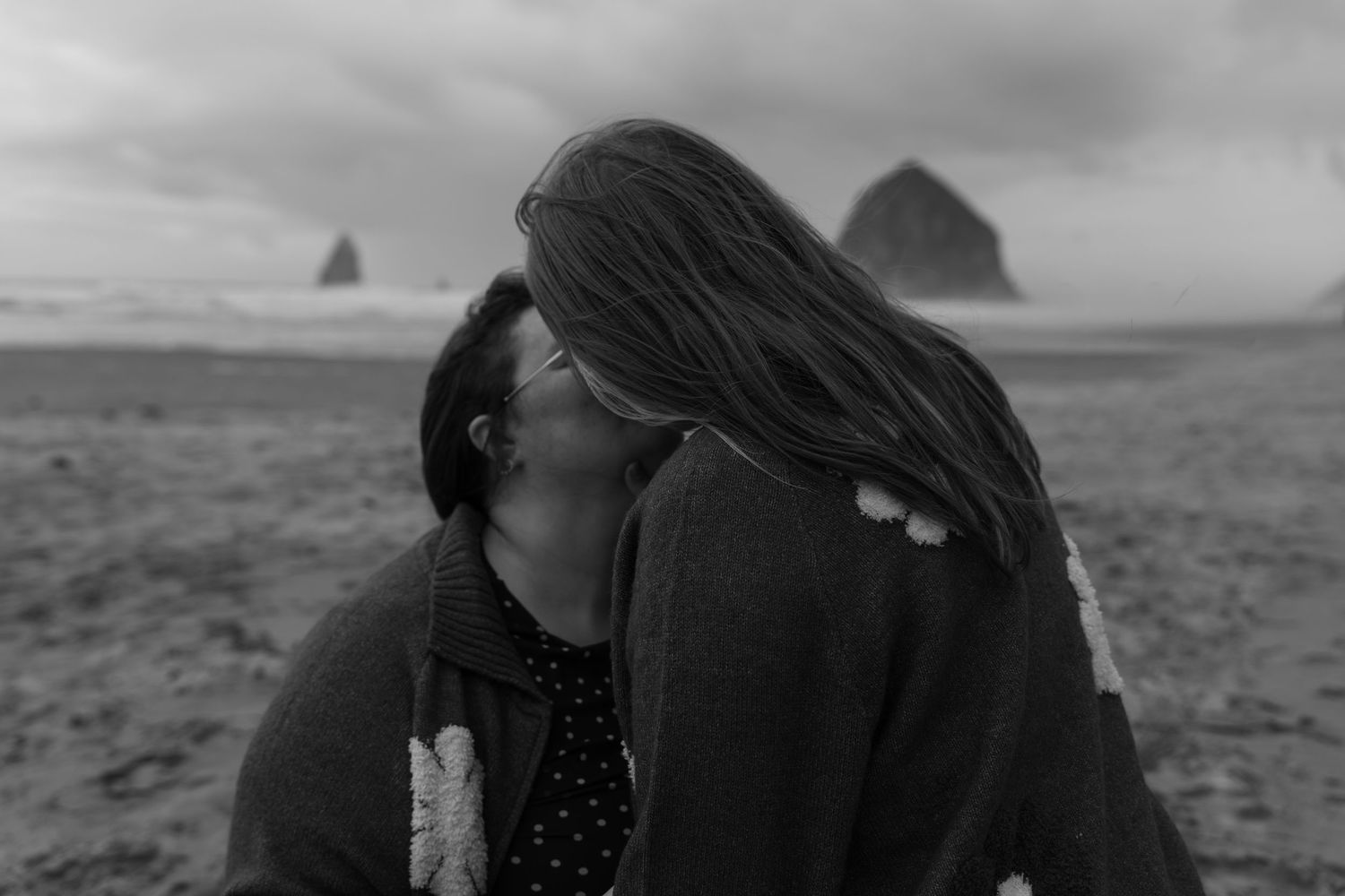 Black and white photo of a couple sharing a romantic kiss on a beach with Haystack Rock in the background.