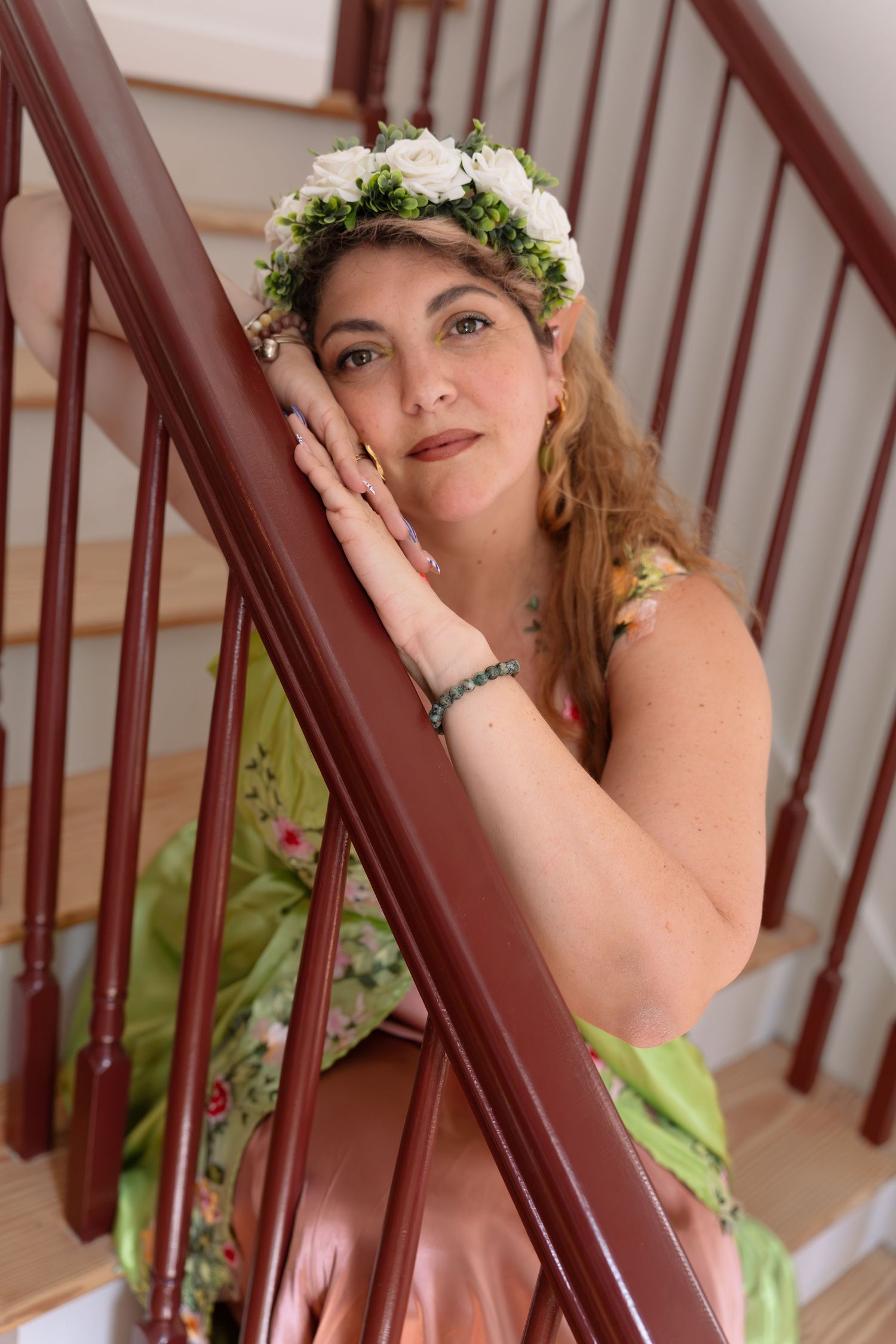 Woman wearing floral crown and green dress poses gracefully on wooden staircase railing.