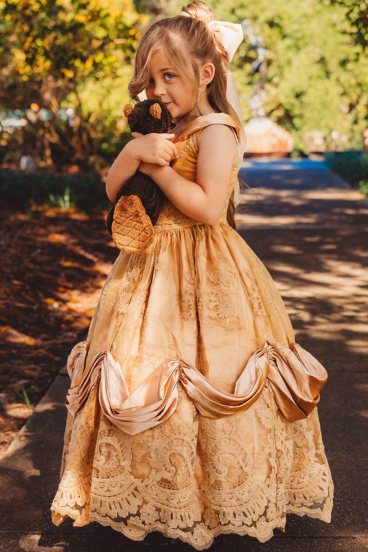 Young girl in golden Belle princess gown holding a stuffed bear toy outdoors in sunlight.