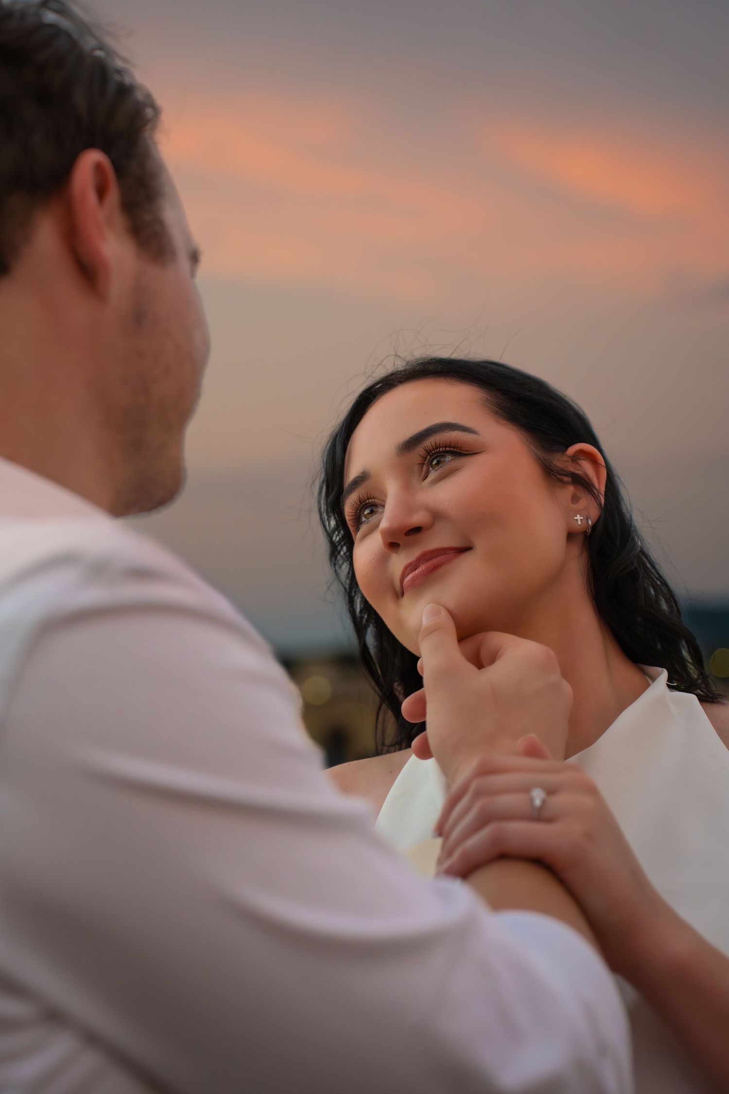 A smiling woman with a ring on her finger gazes lovingly at her partner as he touches her chin during a romantic sunset.