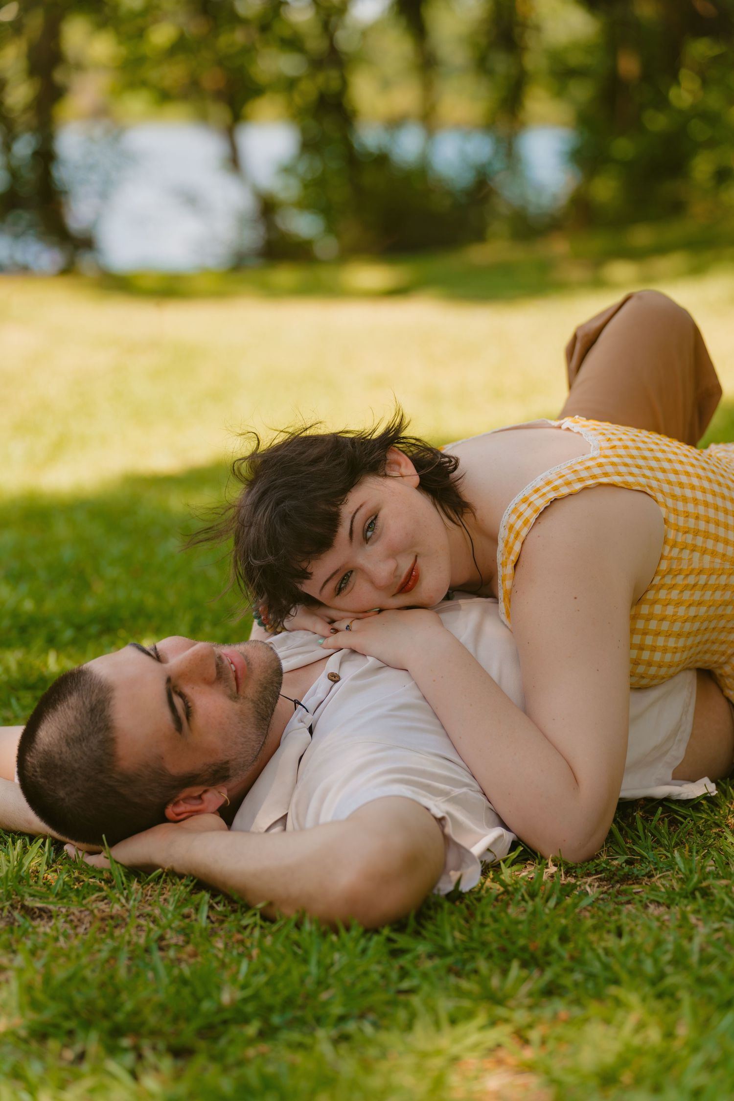 Couple lying on green grass in a park, woman in yellow dress resting on man's chest, sharing a romantic moment outdoors.