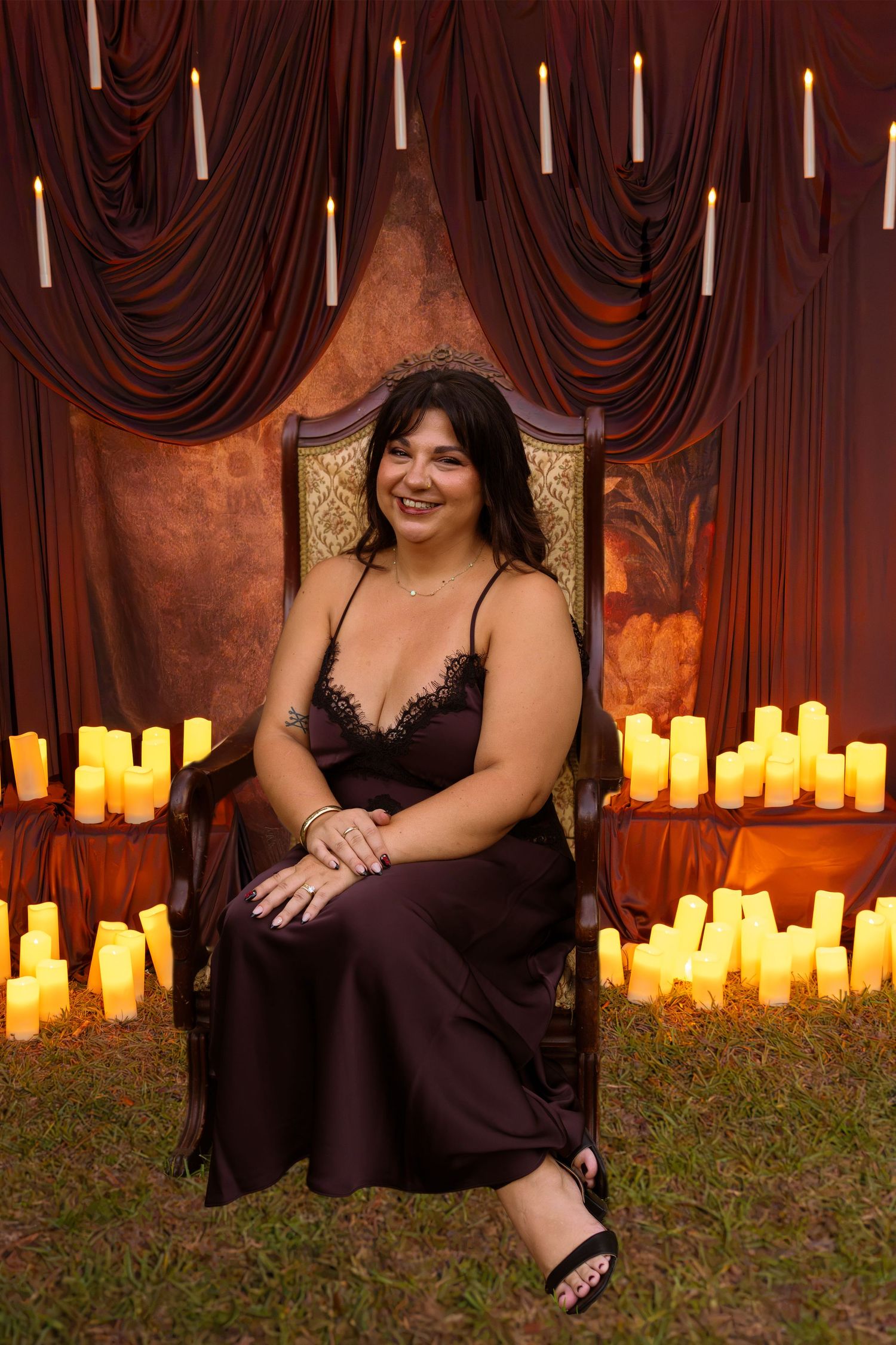 Smiling woman in a dark satin slip dress sits on an ornate chair surrounded by glowing candles and draped burgundy curtains.