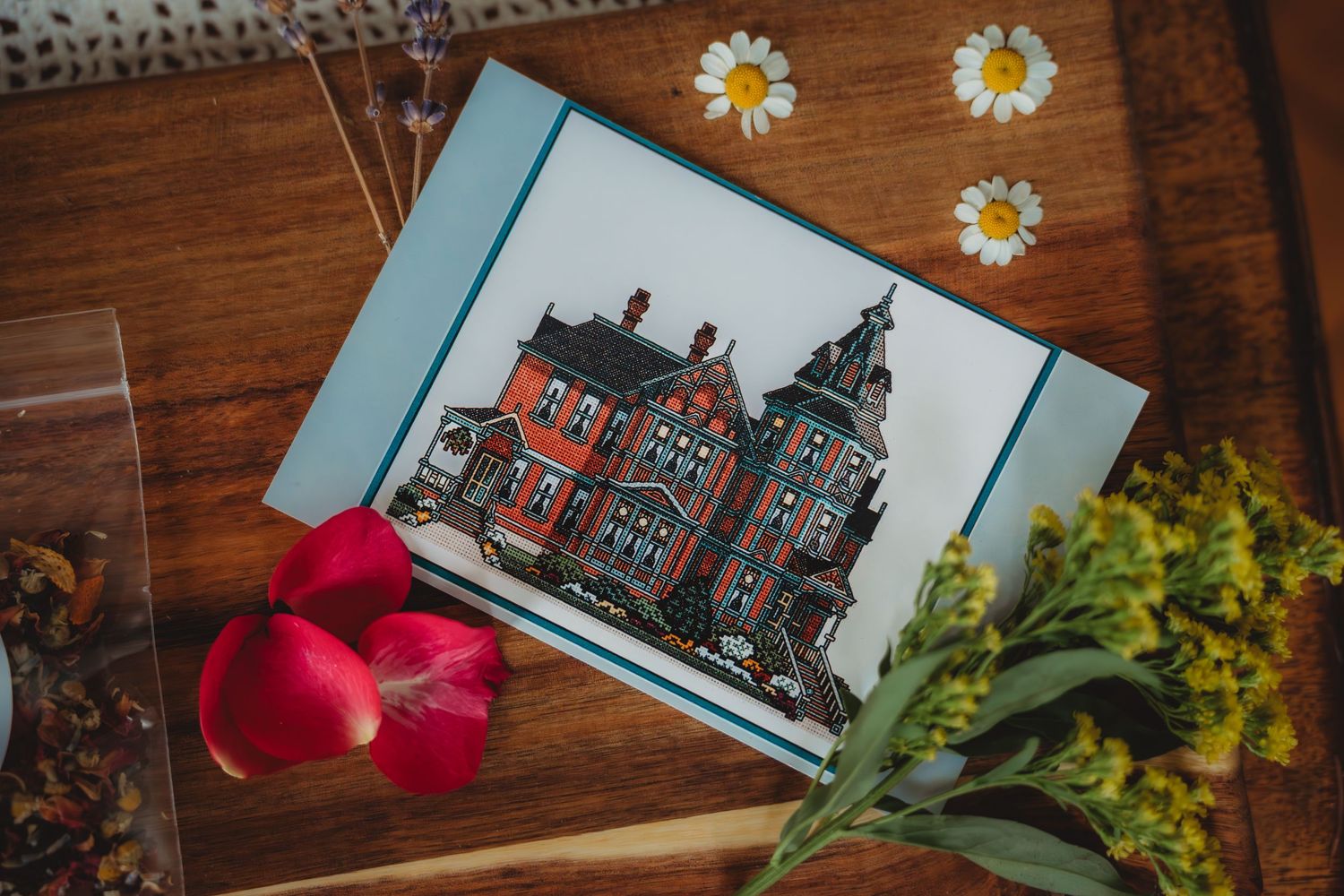 Illustrated greeting card featuring a Victorian mansion on a wooden table with daisies, red petals, and wildflowers.