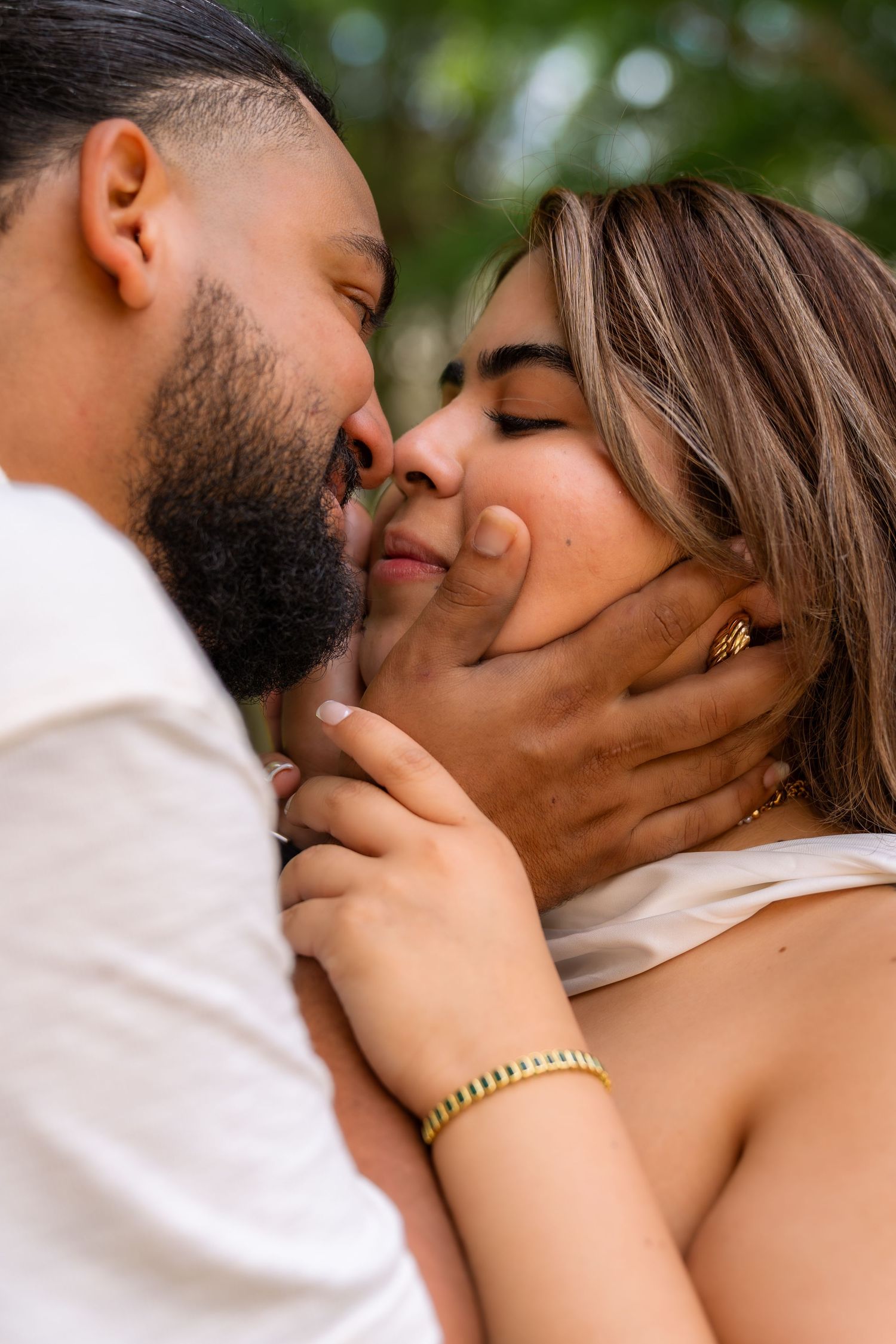 A couple shares a tender kiss outdoors, with the man gently cupping the woman's face, both wearing gold jewelry.