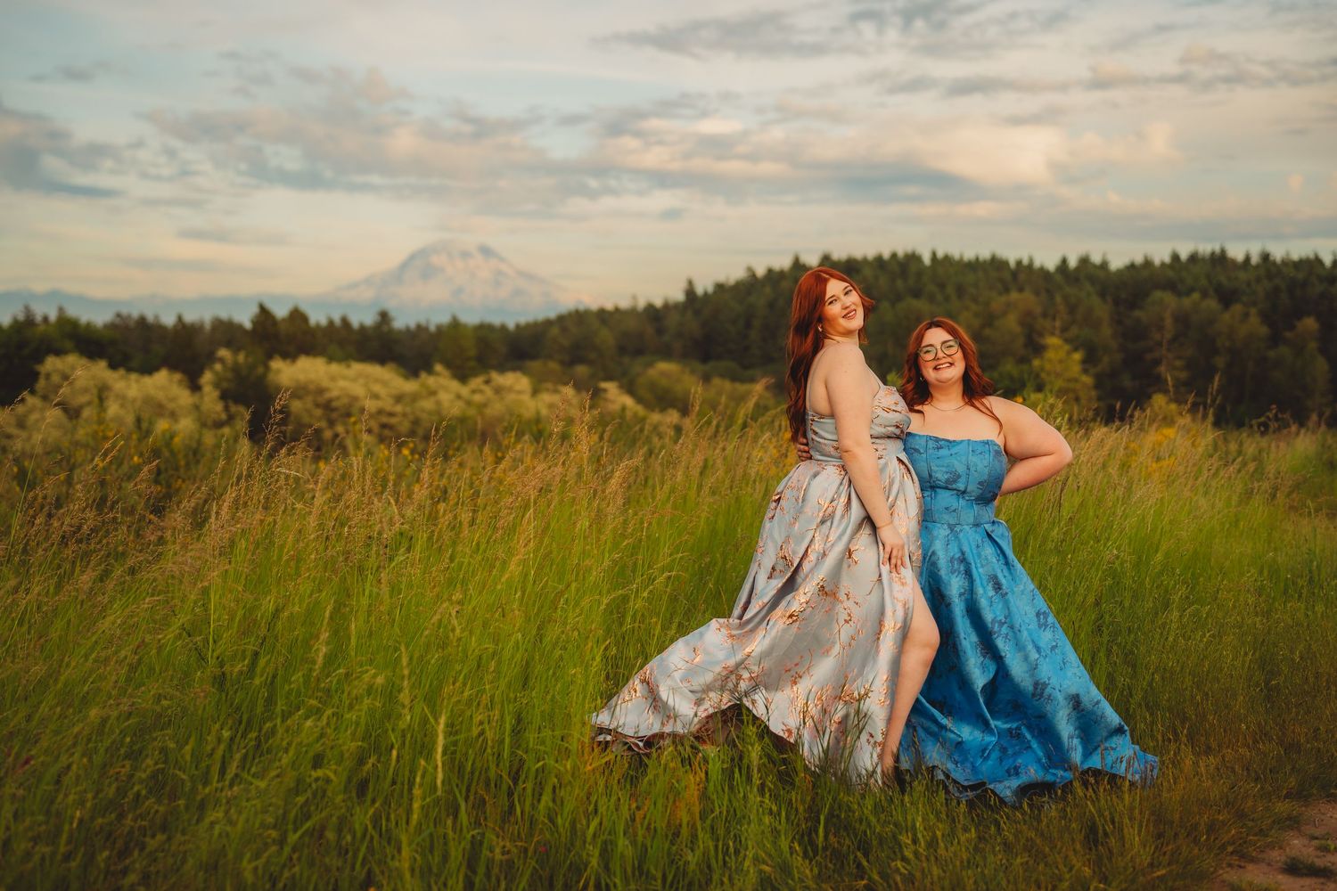 Two women in elegant gowns pose in a lush green meadow with Mount Rainier in the background at golden hour.
