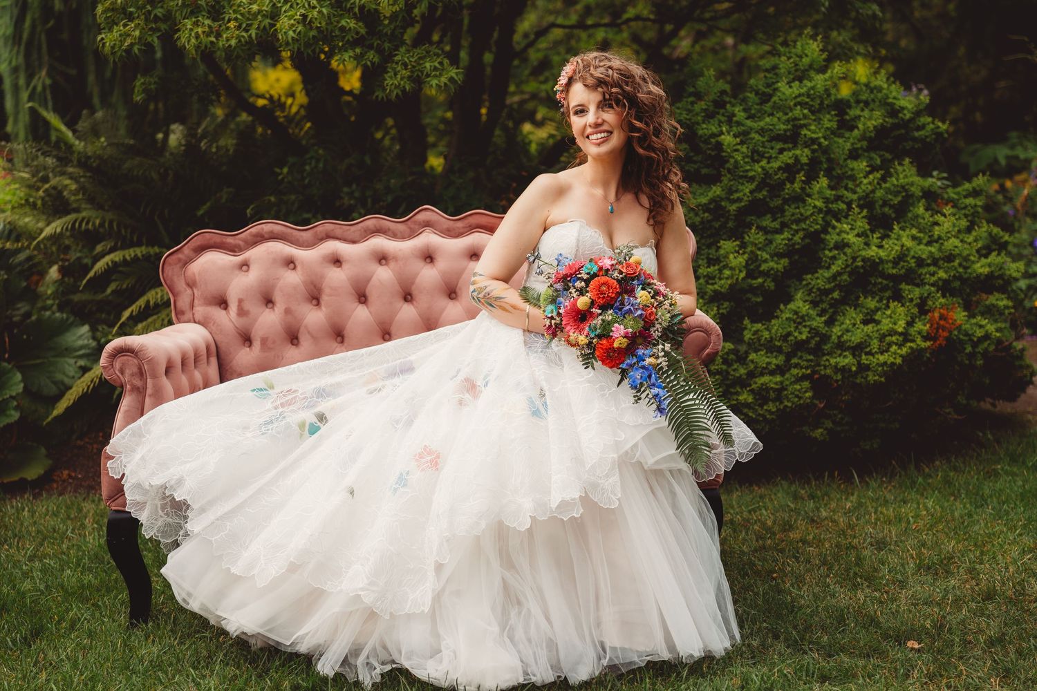 Bride in floral white gown holding colorful bouquet, seated on pink velvet sofa outdoors.
