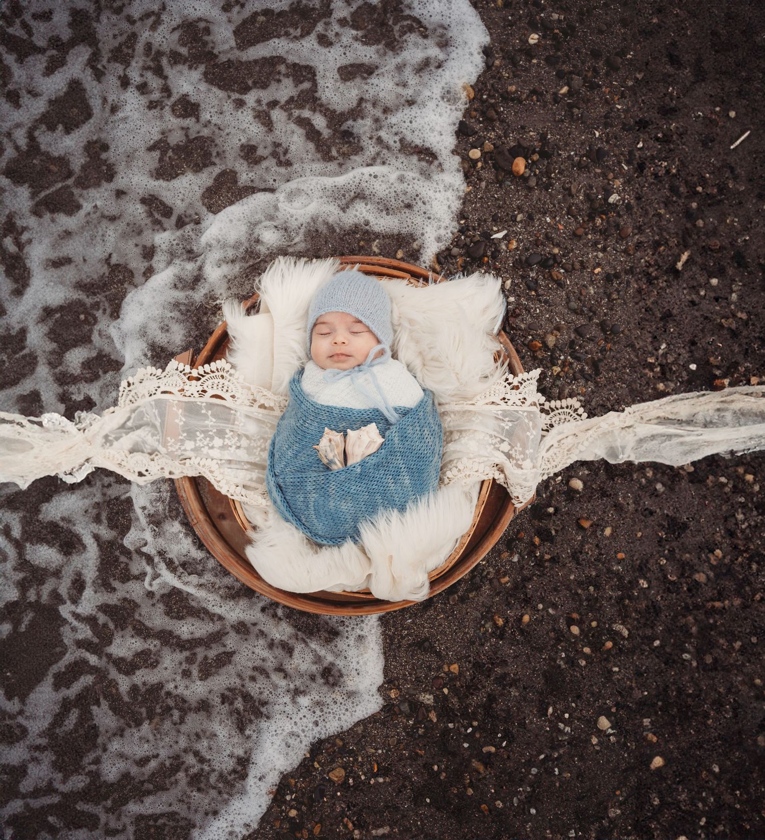 Newborn baby in blue knit outfit sleeping in wicker basket surrounded by ocean waves on beach.