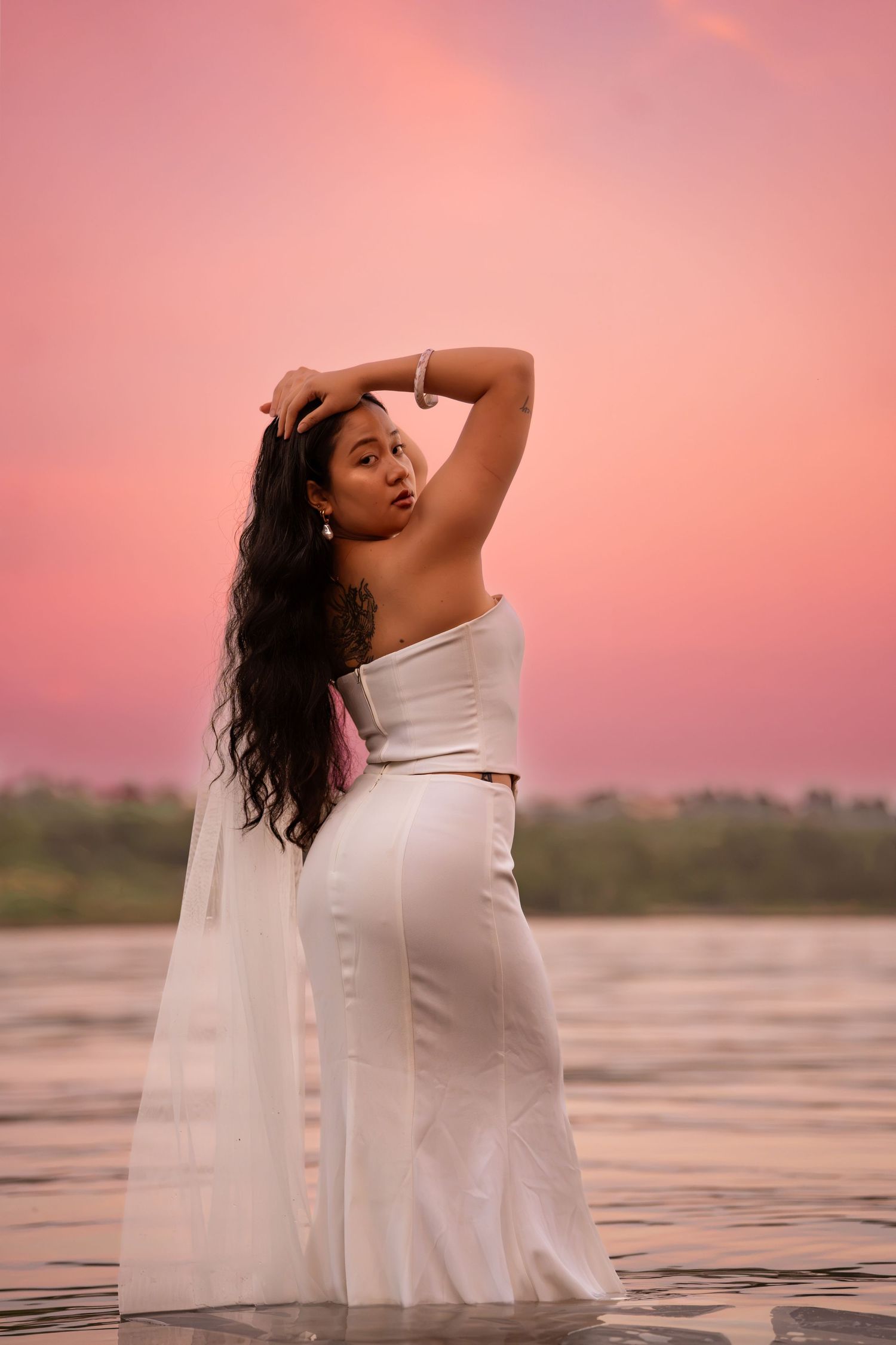 Woman in white strapless top and skirt poses gracefully by water under a stunning pink sunset sky.