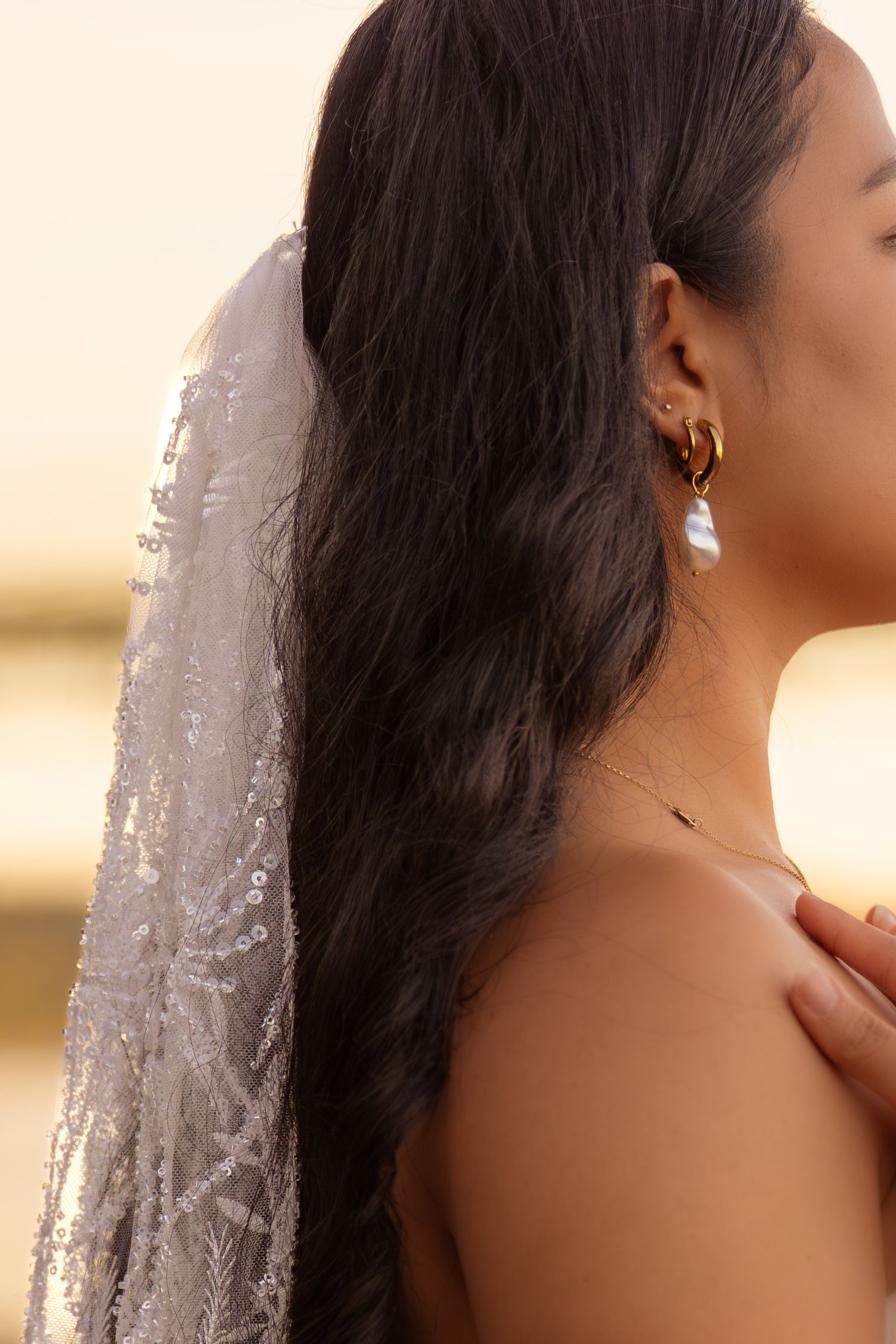 Bride with dark hair wearing a sequined veil and gold pearl drop earrings at golden hour.
