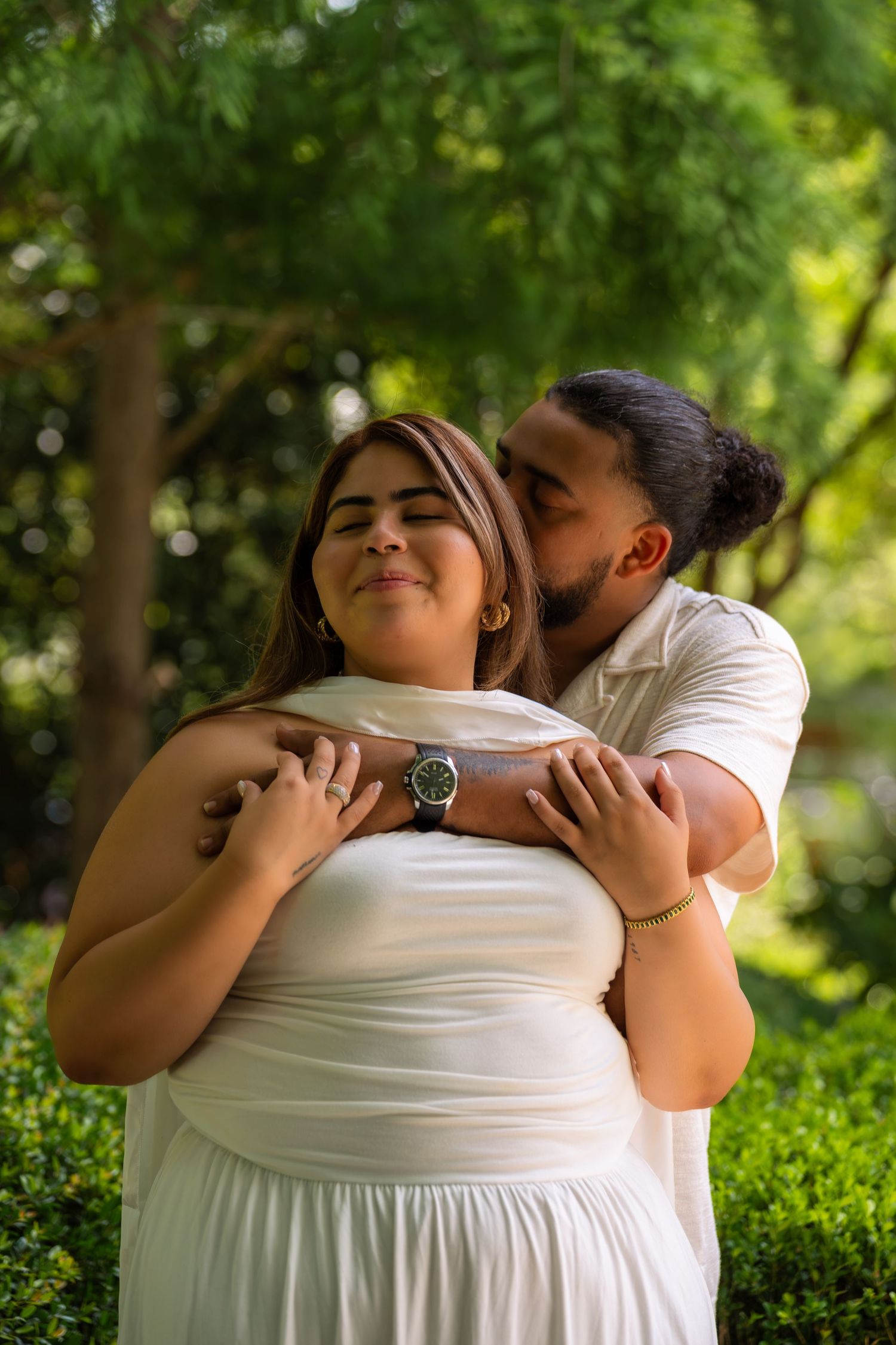 A couple embraces outdoors, man kissing woman's cheek, both in white attire, surrounded by lush green trees.