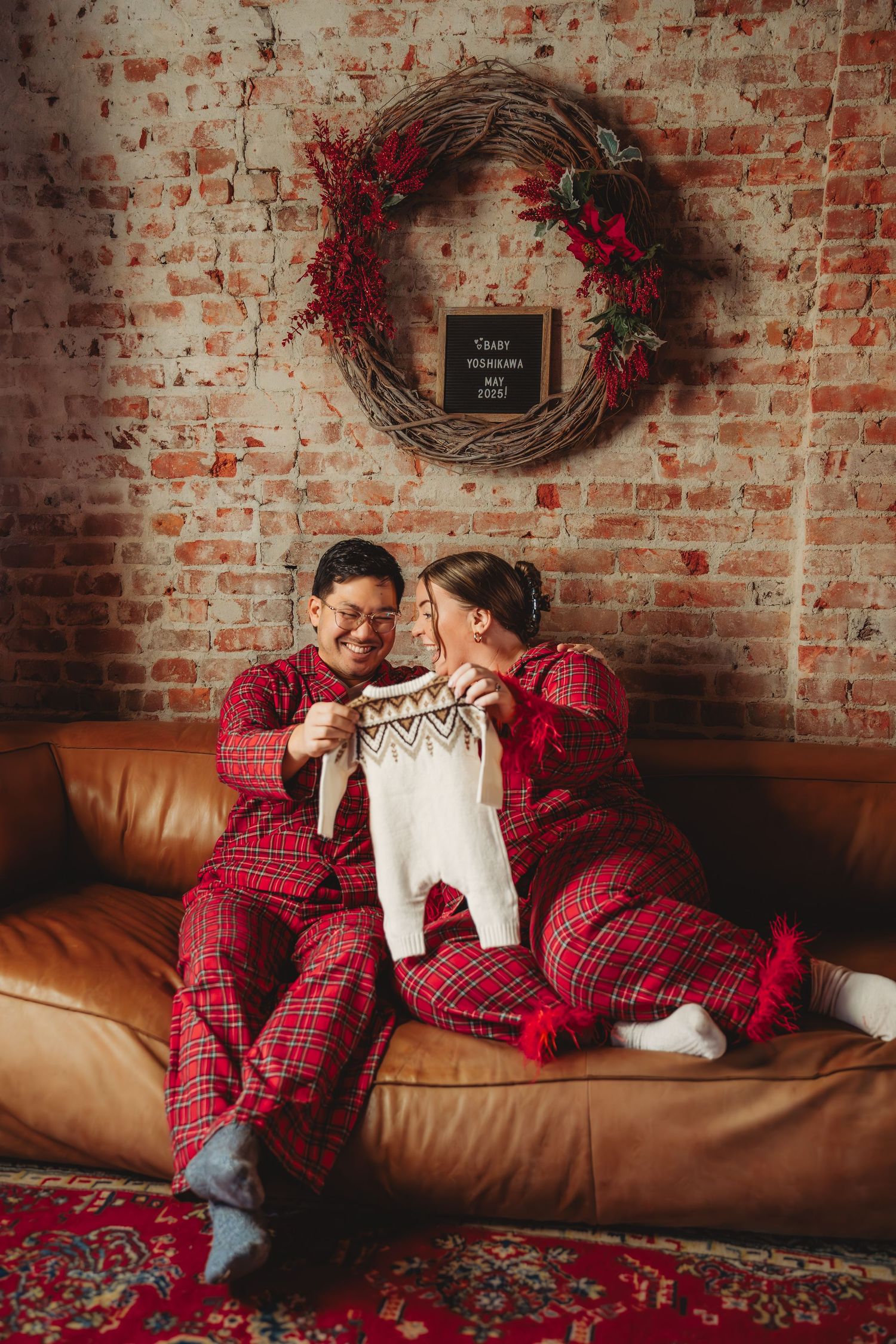 Couple in matching red plaid pajamas holding baby clothes on a couch with Christmas wreath. Seattle